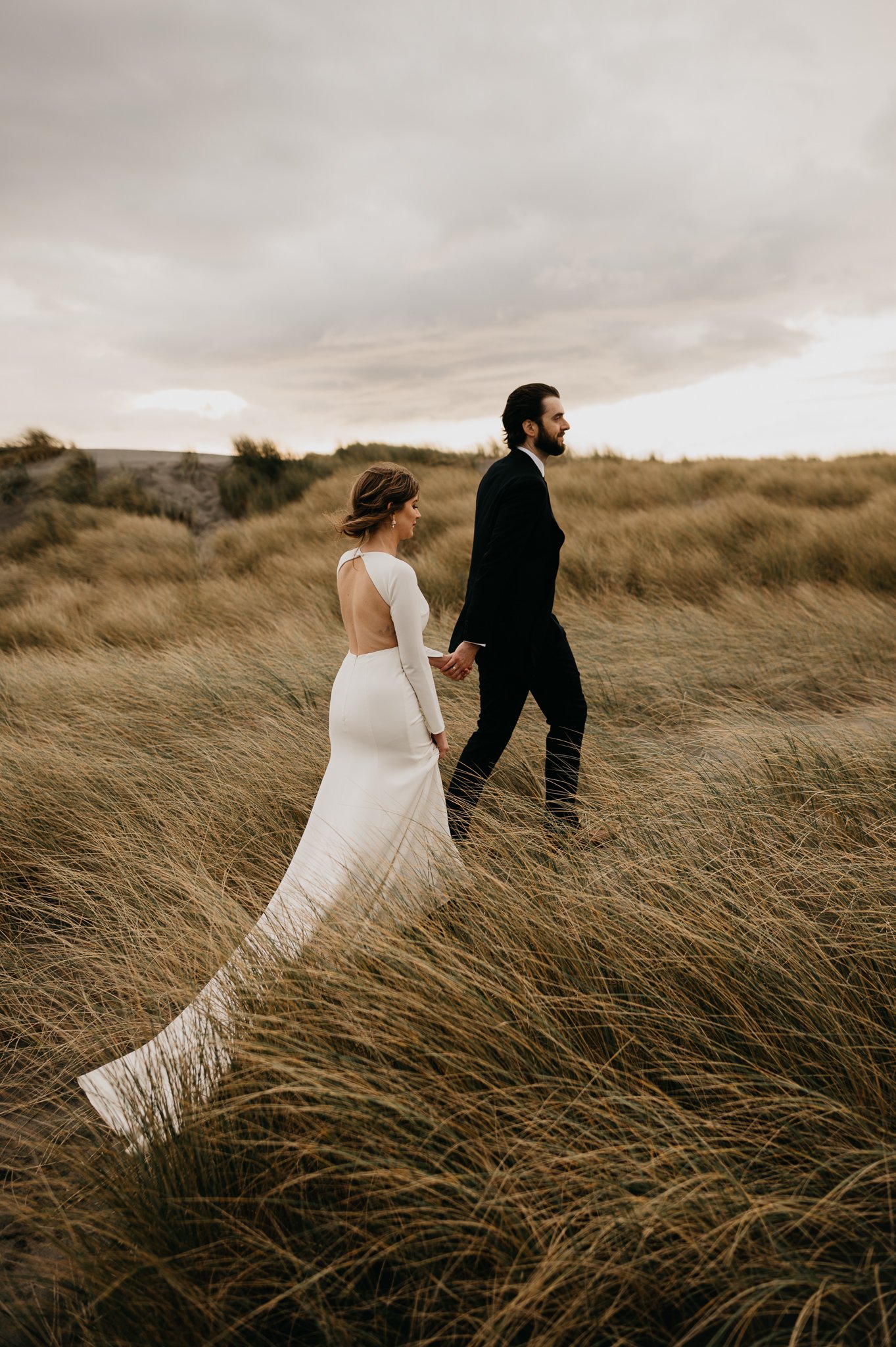 Bride and groom in wedding attire walking hand and hand in the tall Oregon seagrass.