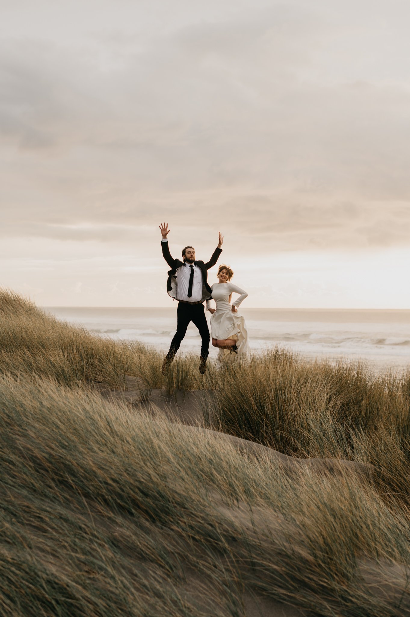 Bride and groom in wedding attire on sand dune with tall sea grass around them jumping up in the air with joy.