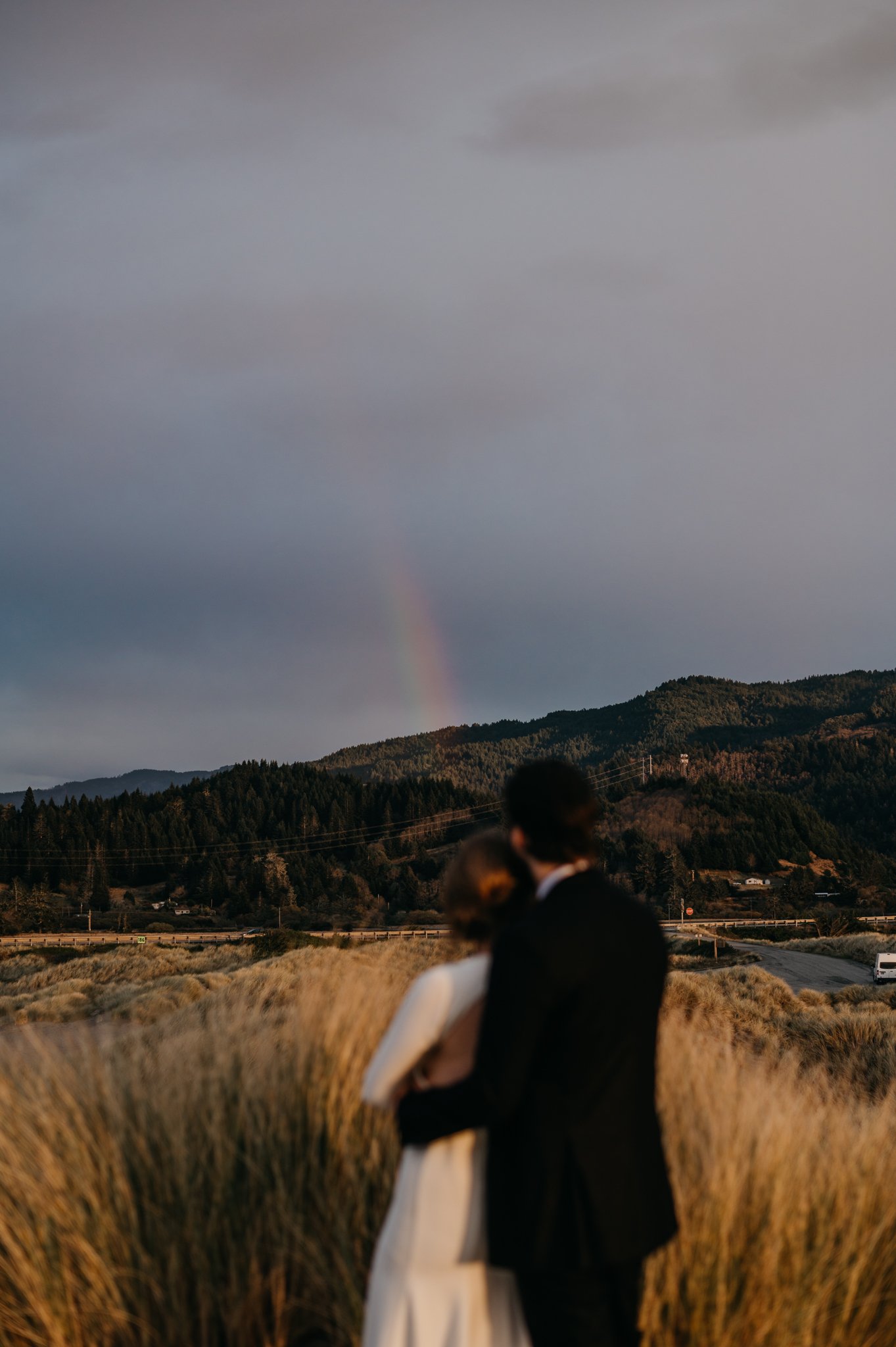 Bride and groom standing on beach with a moody grey sky and a rainbow in the Oregon sky.