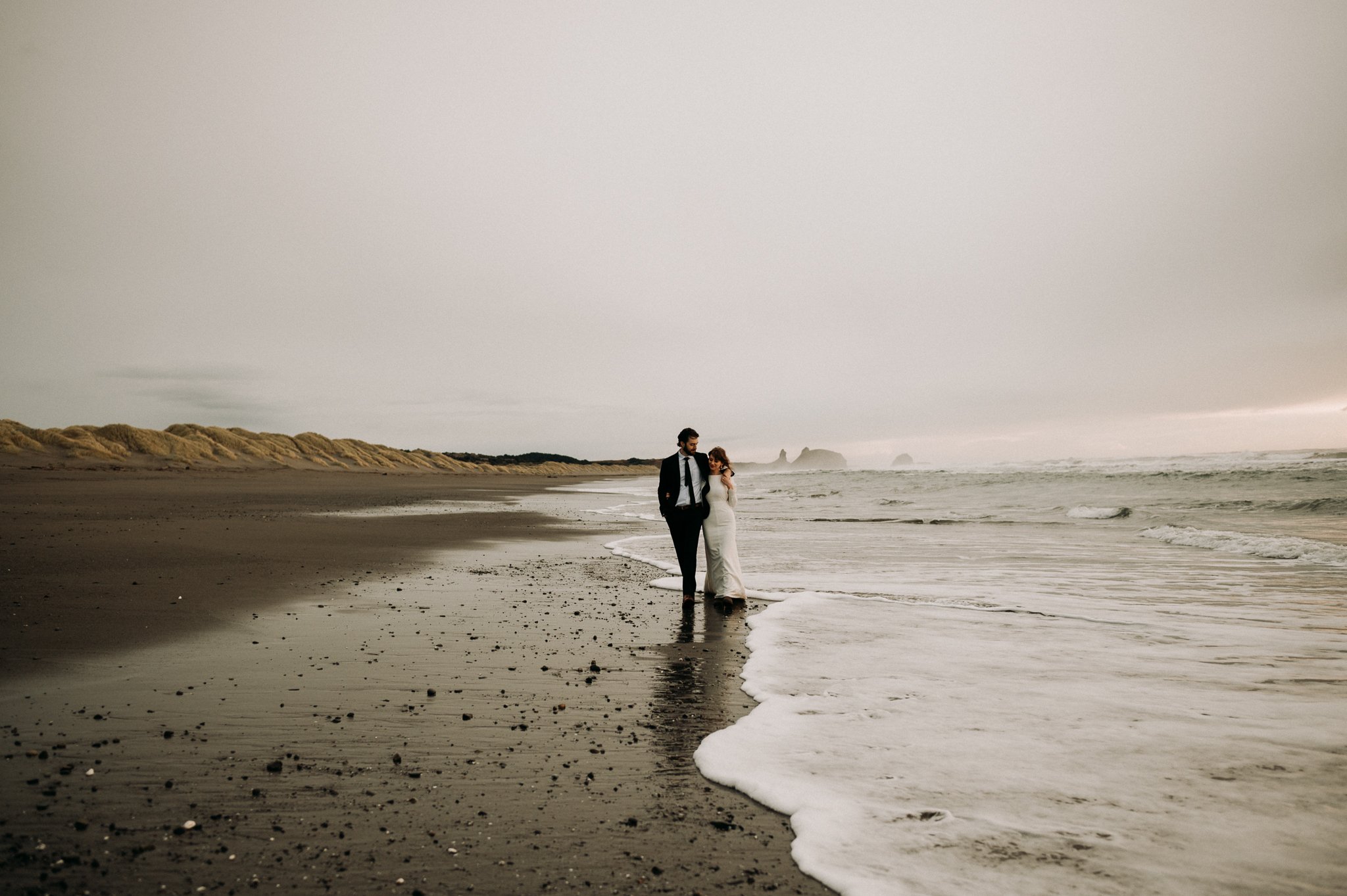 Bride and groom in wedding attire walking along the shore break after wedding ceremony.
