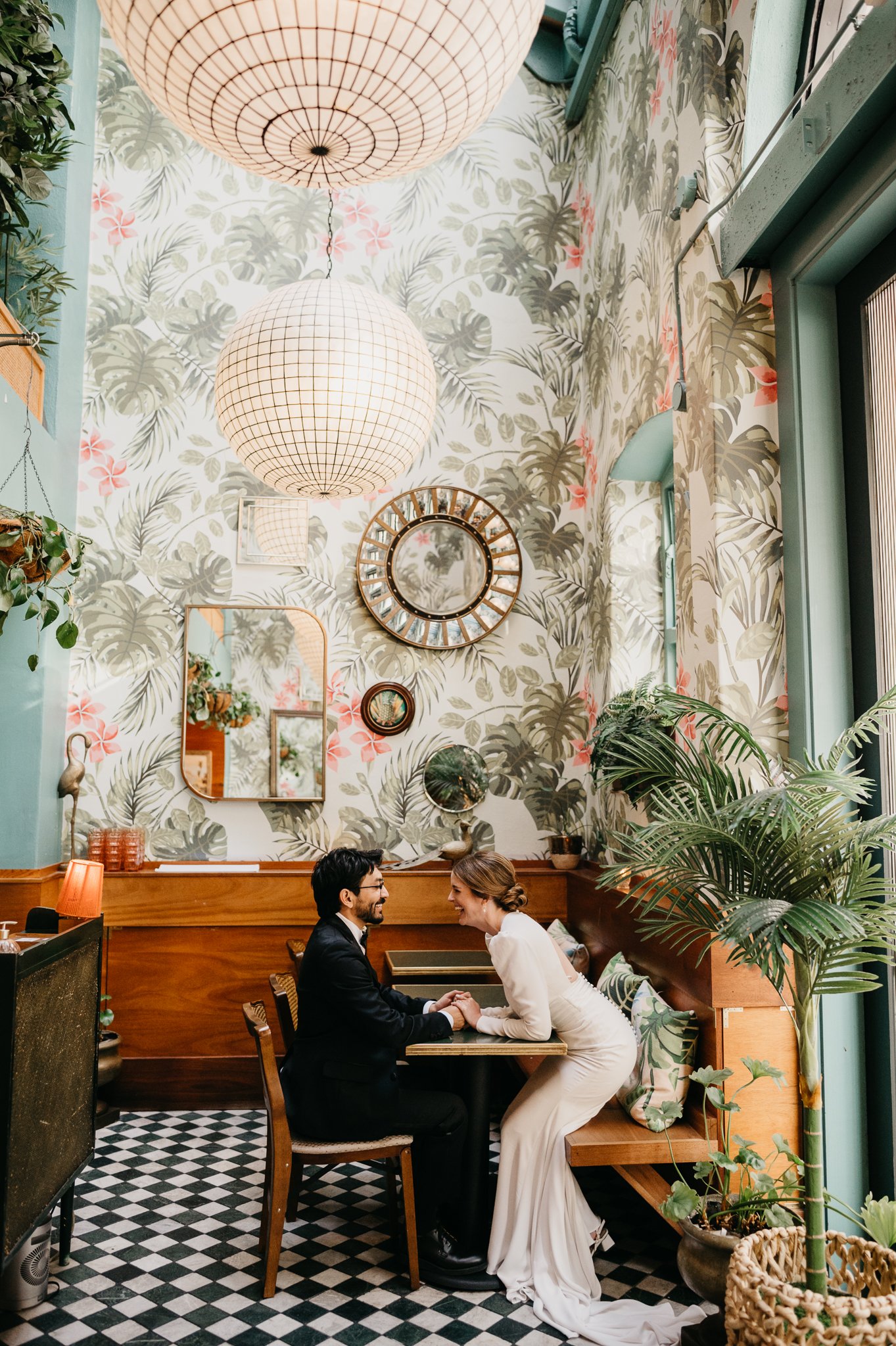 Couple dressed formally, sitting at a table in a colorful restaurant with tropical decor, holding hands and smiling at each other.