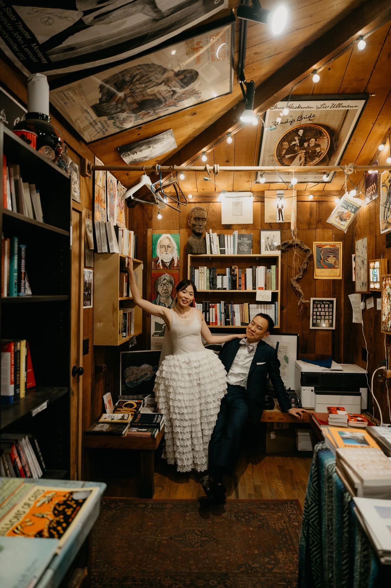 A woman in a white dress and a man in a suit are inside a cozy, wood-paneled bookstore, with bookshelves filled with books and artwork on the walls, surrounded by string lights.