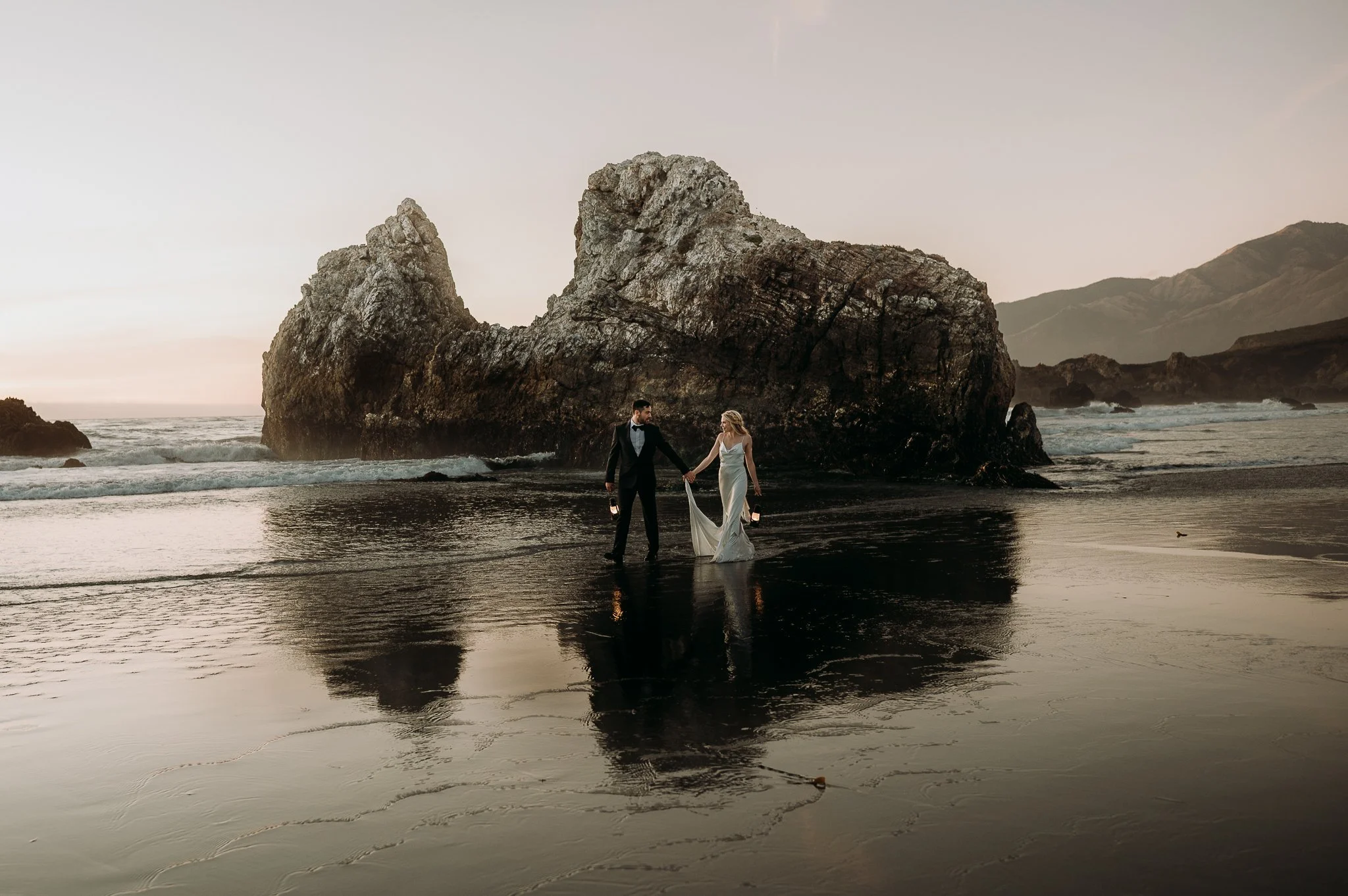 A couple dressed in wedding attire holding hands on a beach at sunset, with a large rock formation and mountains in the background.