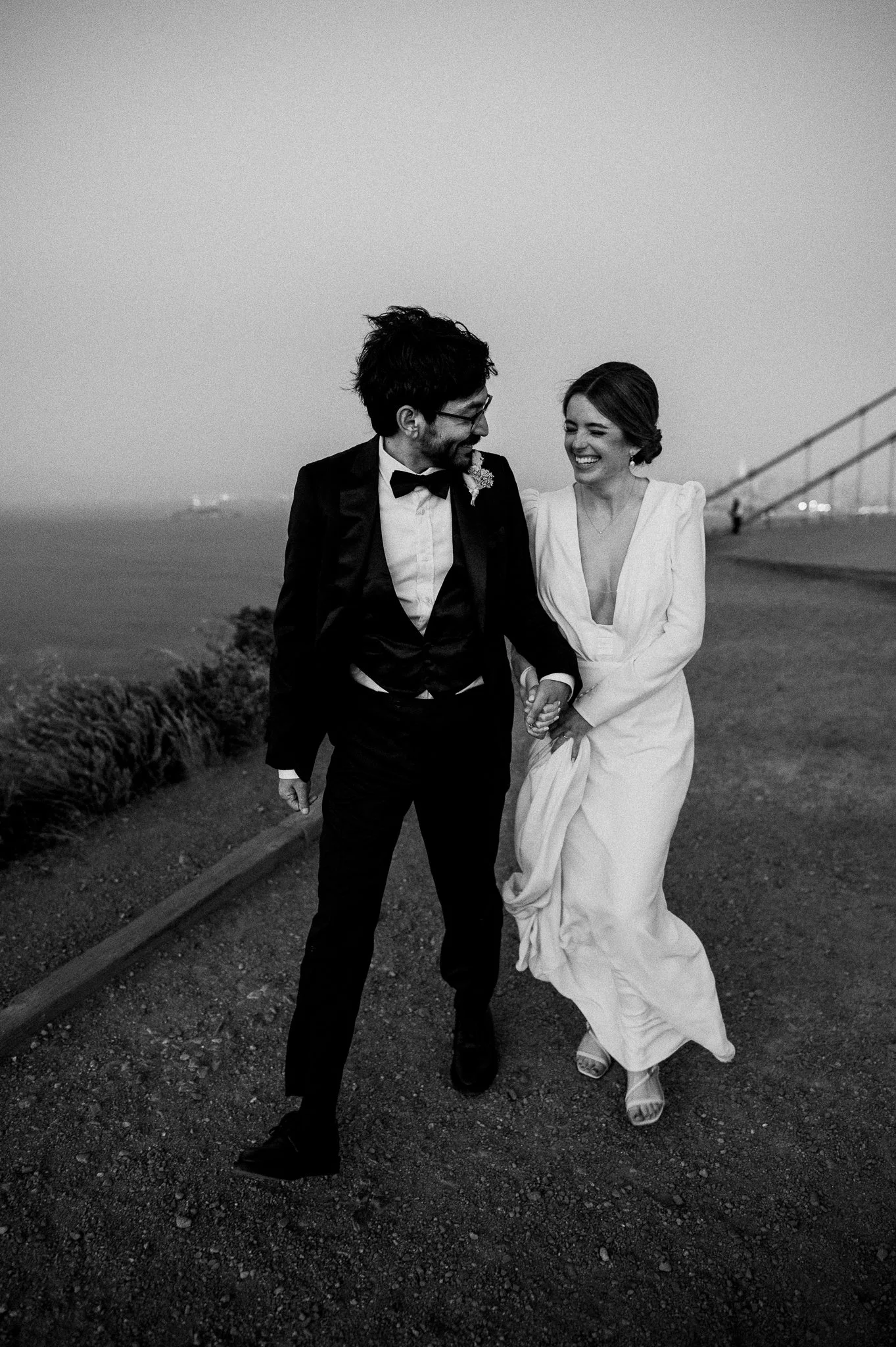 A black and white photo of a newlywed couple walking hand in hand outdoors by the water, smiling and looking at each other, with a bridge in the background.