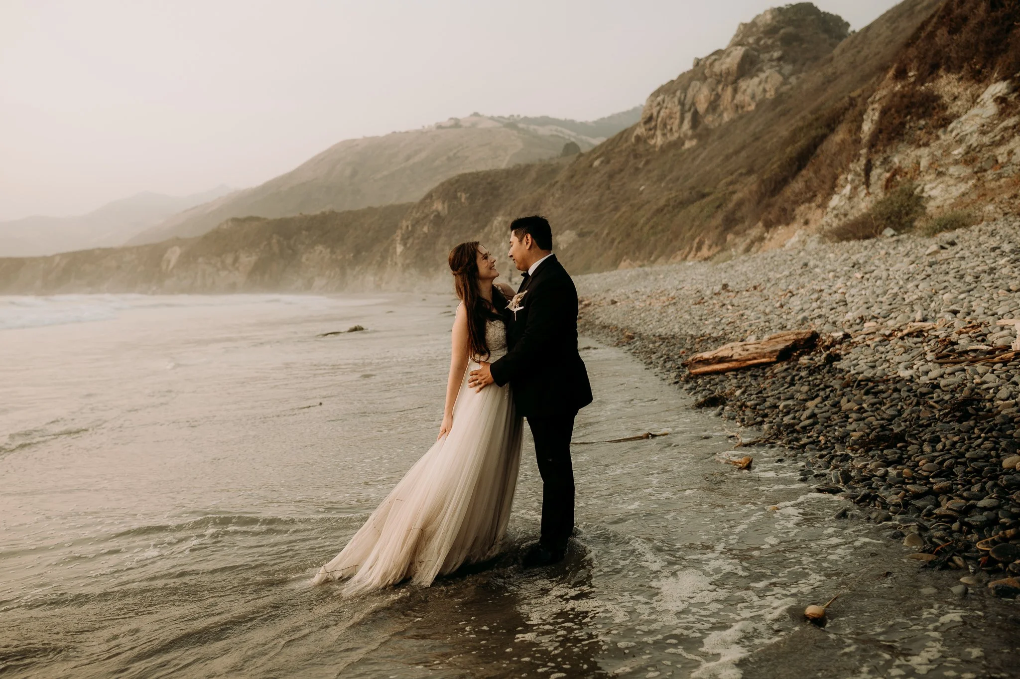 A bride and groom standing in the shallow water at the beach, looking at each other lovingly with mountains in the background.