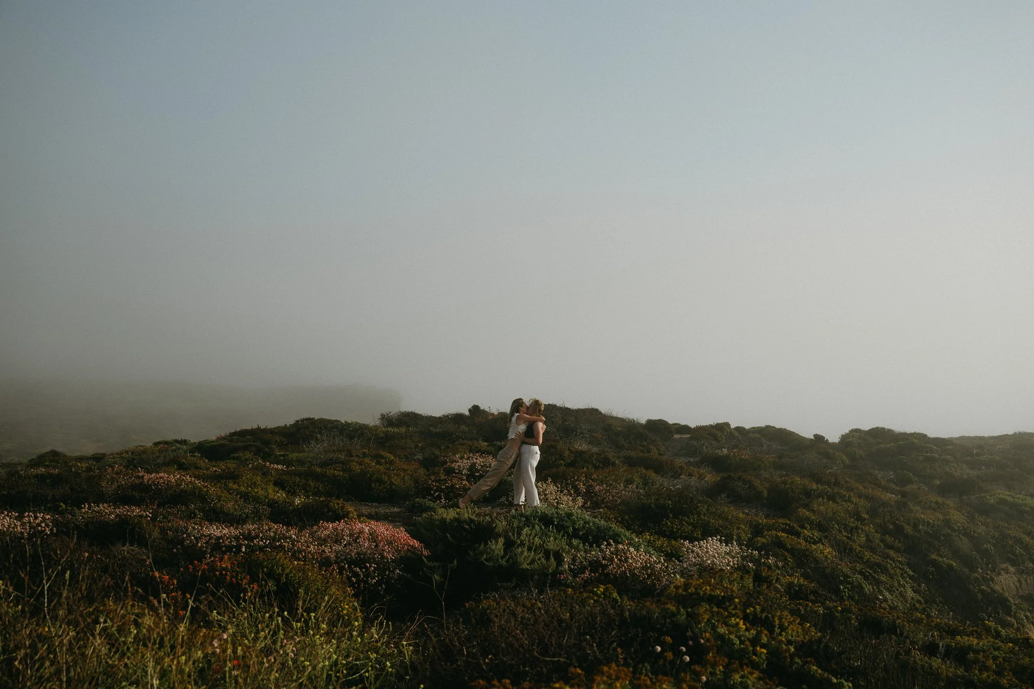 Two people hugging on a grassy hilltop with bushes, fog, and overcast sky.