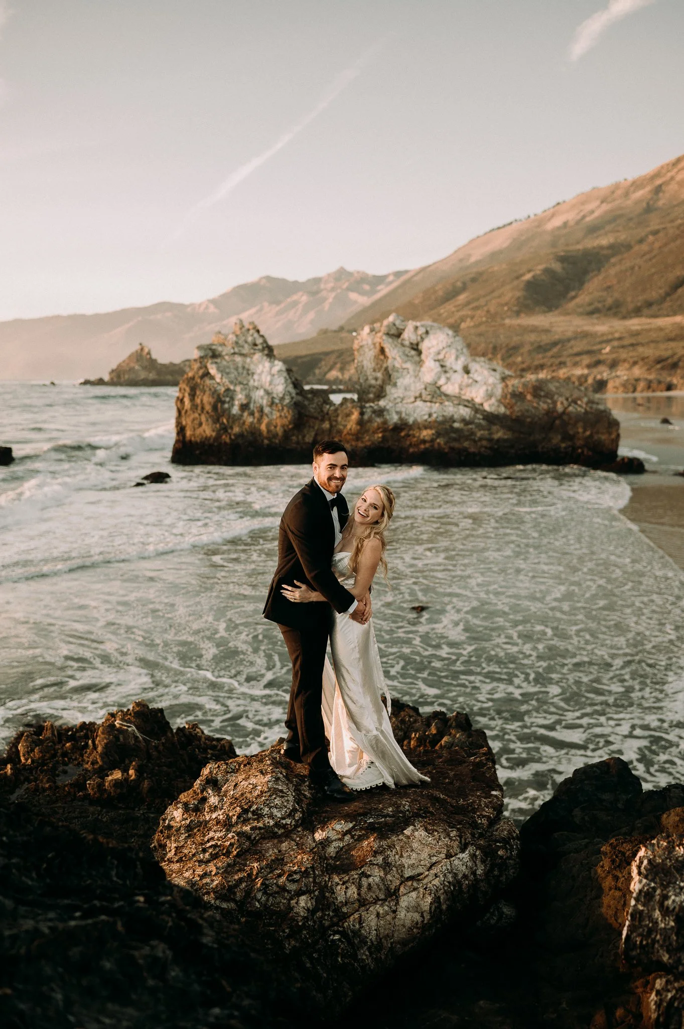 A happy couple in wedding attire standing on a rocky beach at sunset, with mountains and large rock formations in the background.