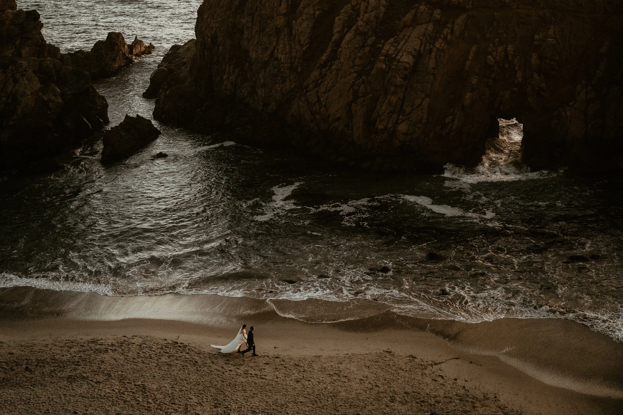 A bride and groom walking on a beach near a large rock formation with a natural archway, ocean waves, and cliffs