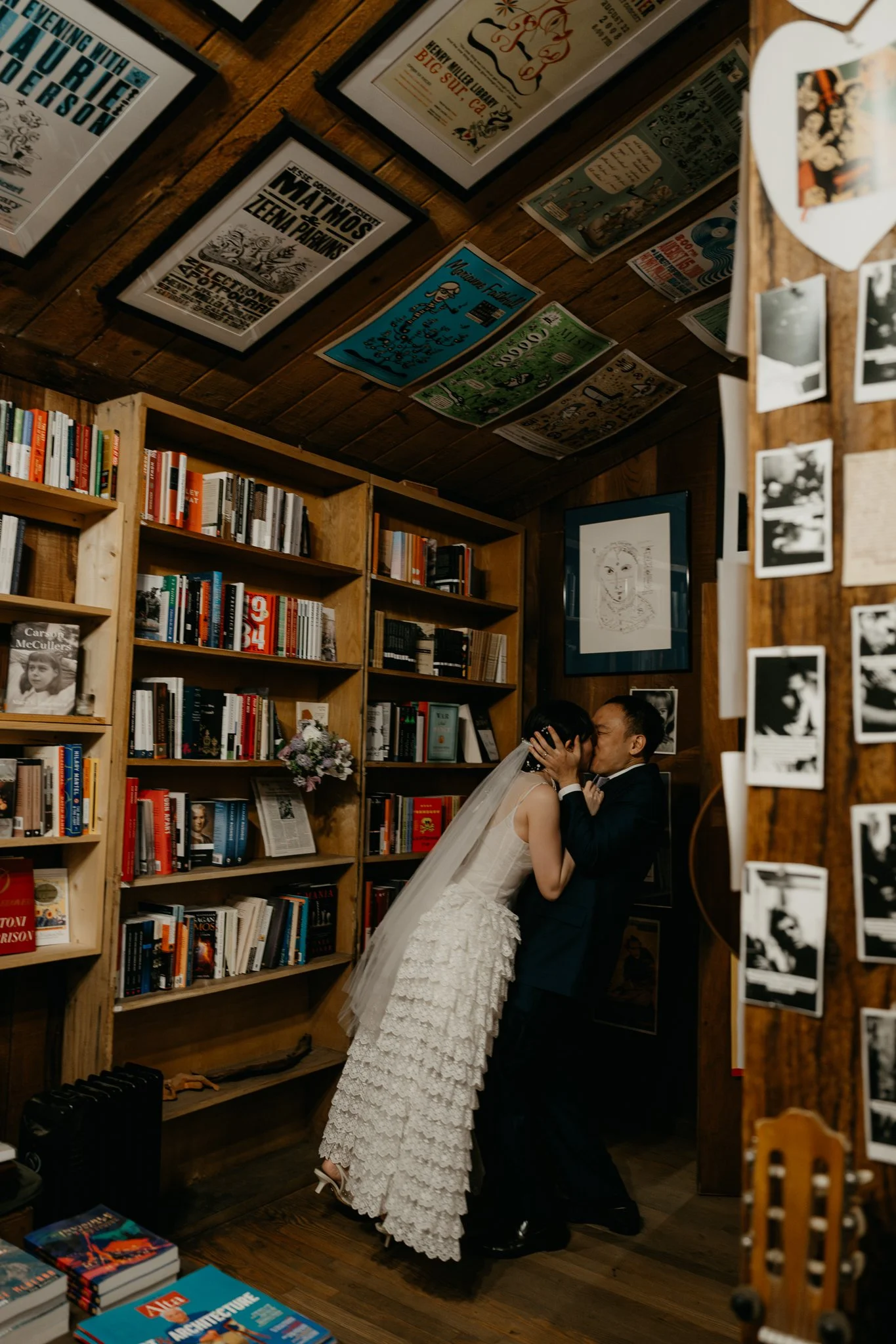 A bride and groom sharing a kiss in a cozy, wood-paneled room decorated with books and artwork.