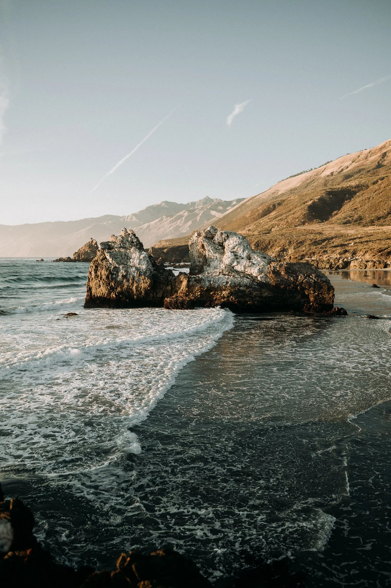 A scenic coastal landscape featuring large rocks in the ocean with waves crashing against them, green hills in the background, and a clear sky with a few clouds.