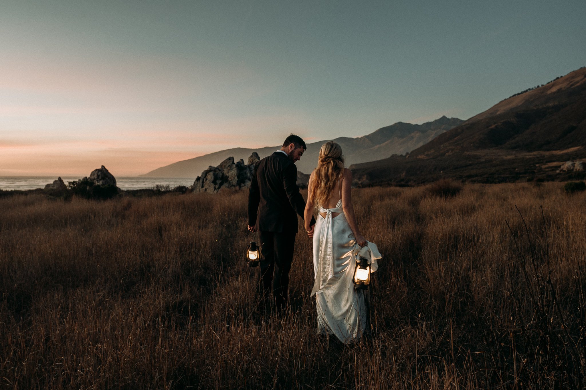 A man and woman in wedding attire walking through a grassy field at sunset, holding lanterns, with mountains and ocean in the background.