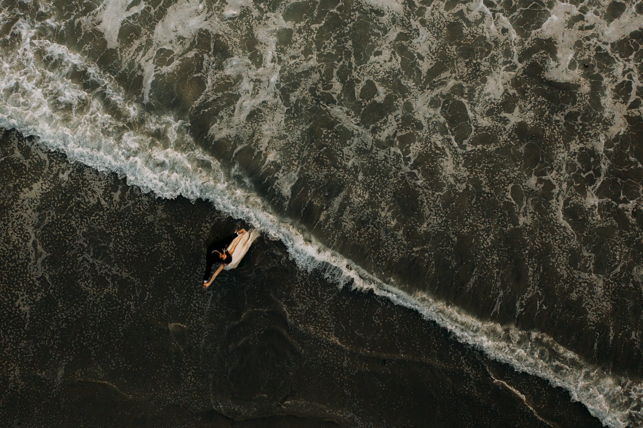 A person lying on a surfboard on the sand at the edge of the ocean with waves approaching.