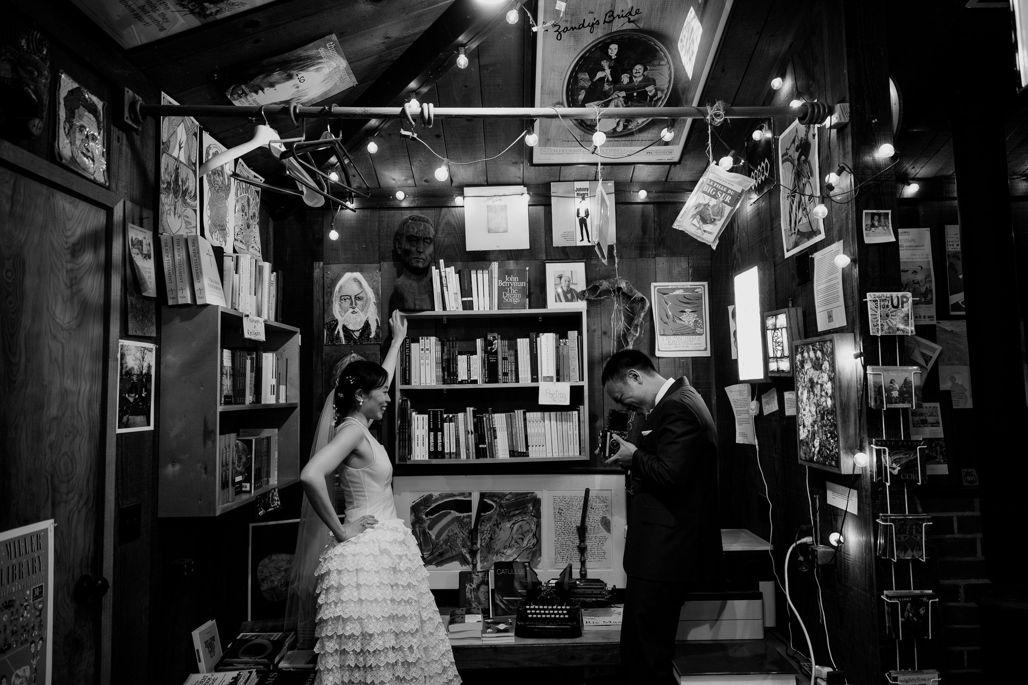 A wedding couple in a bookstore, the bride in a wedding dress smiling and touching her head, the groom in a suit looking at a camera, surrounded by books, artwork, and decorative lights.