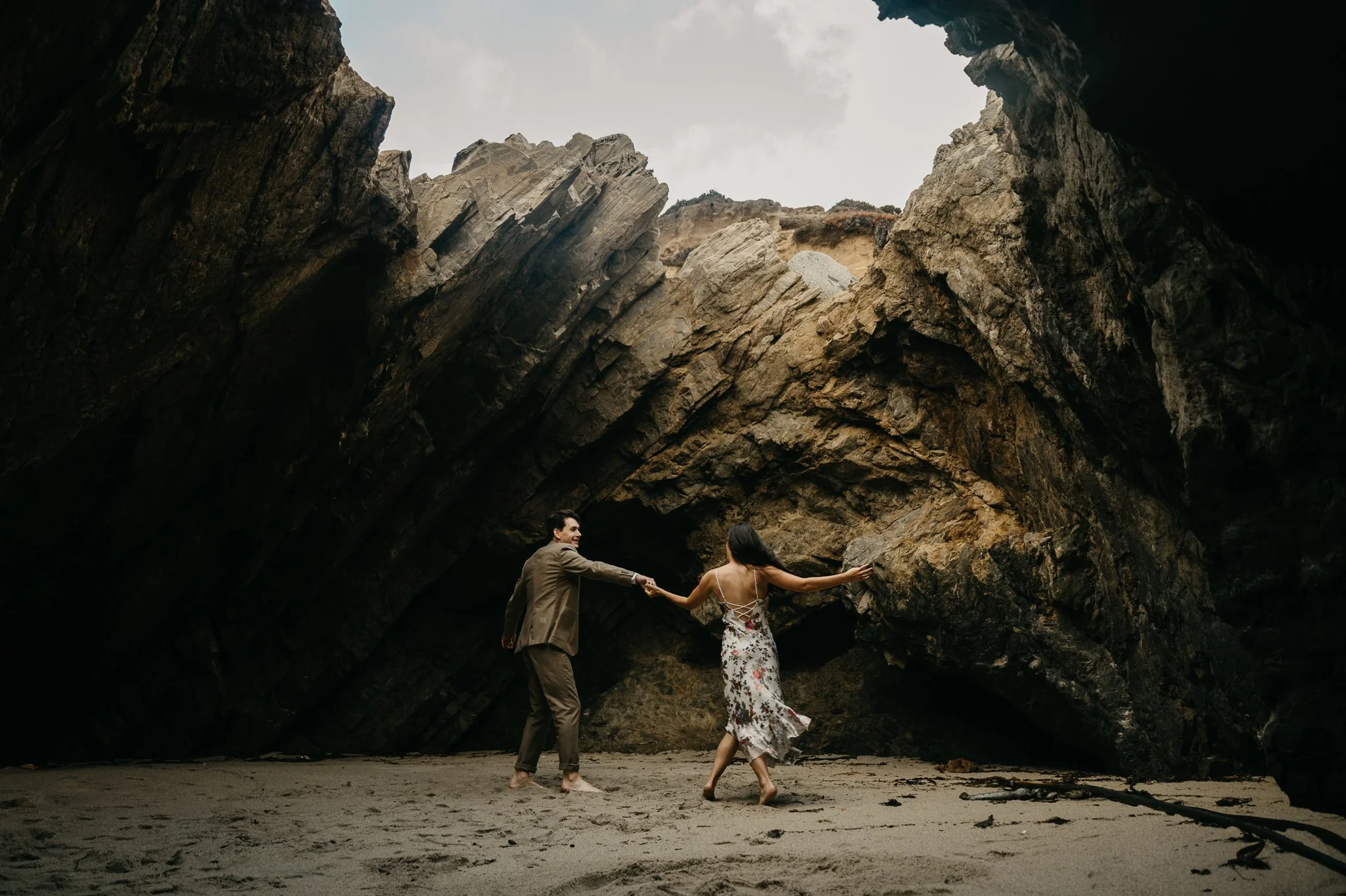 A couple in formal attire dancing on a sandy beach inside a large rock formation with an opening to the sky.