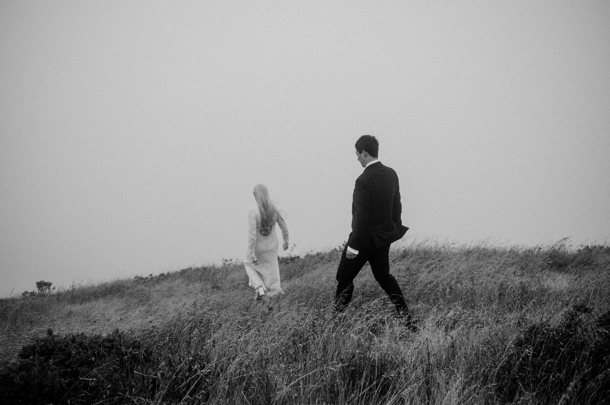 A black-and-white photo of a man in a suit walking behind a woman in a long dress on a grassy hill.