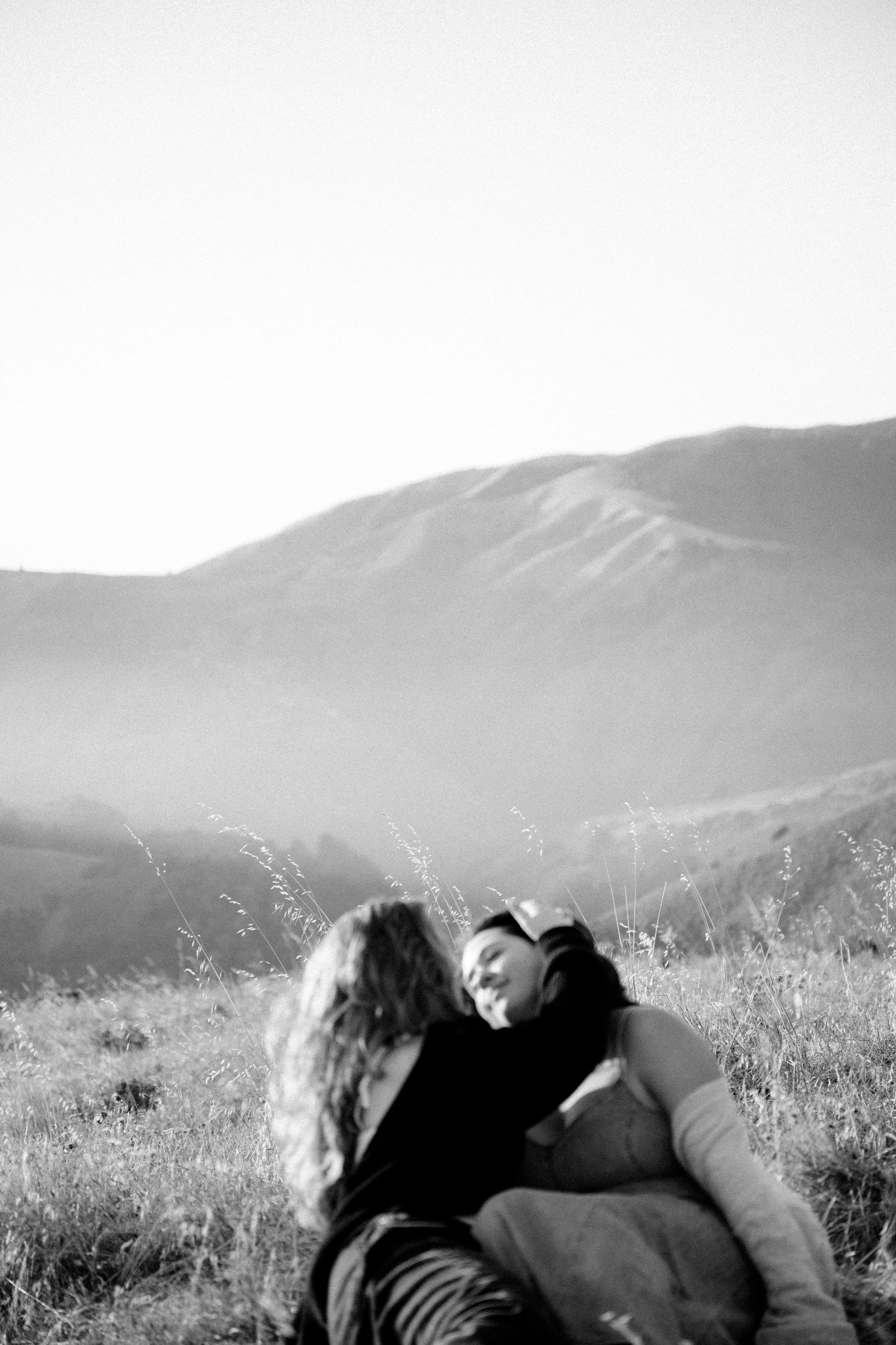 Two women lying on grass in a mountain landscape, embracing and looking at each other.