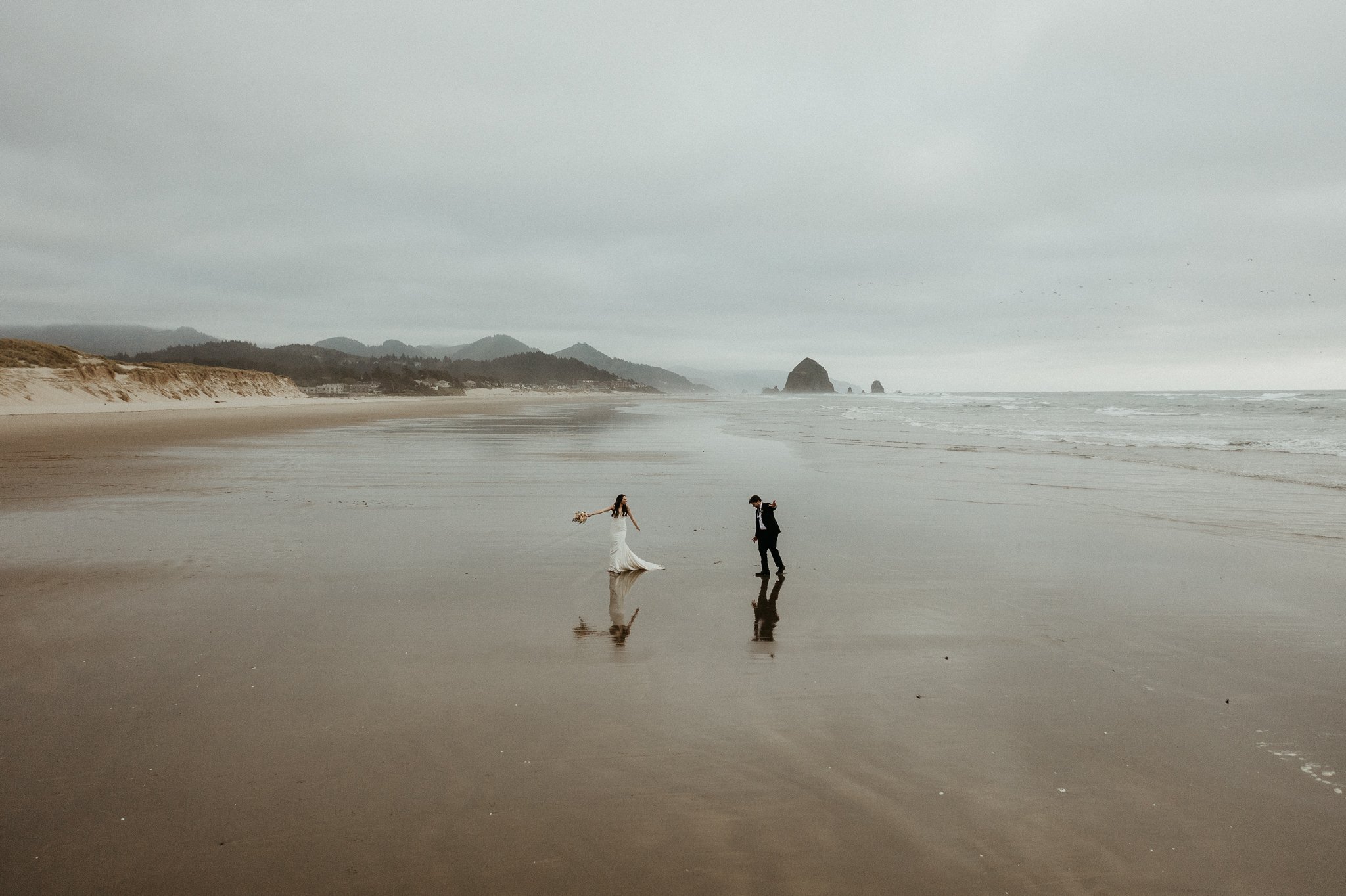 A couple dressed in wedding attire standing on a sandy beach with overcast skies, waves gently lapping at their feet, and distant rock formations and hills in the background.