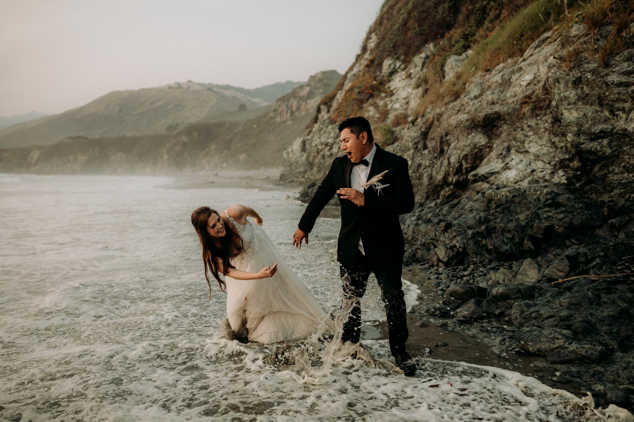 A newlywed couple in formal wedding attire playing and splashing water at the beach with rocky cliffs and hills in the background.