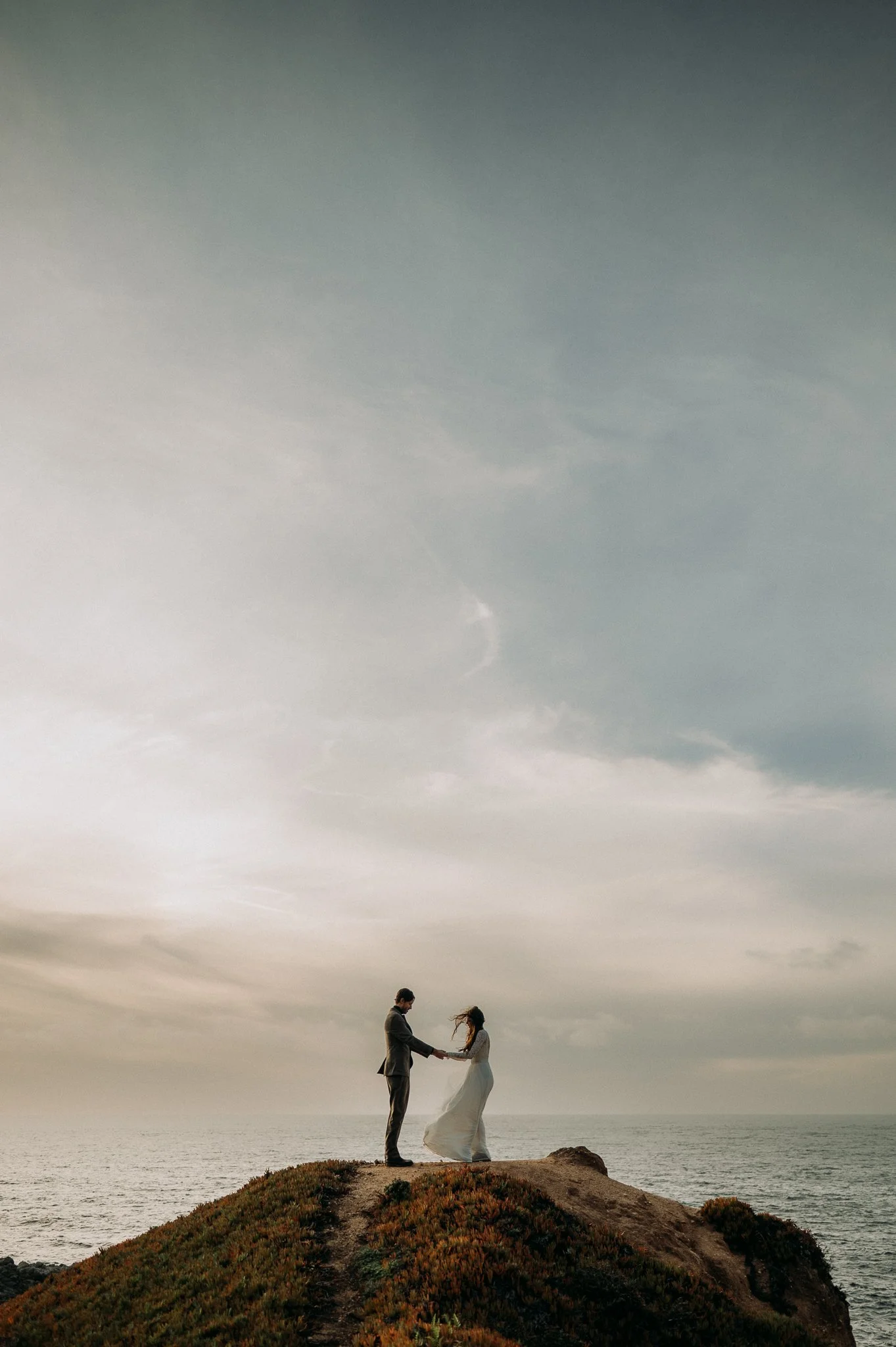 A couple in formal attire exchanging vows on a hilltop overlooking the ocean at sunset.