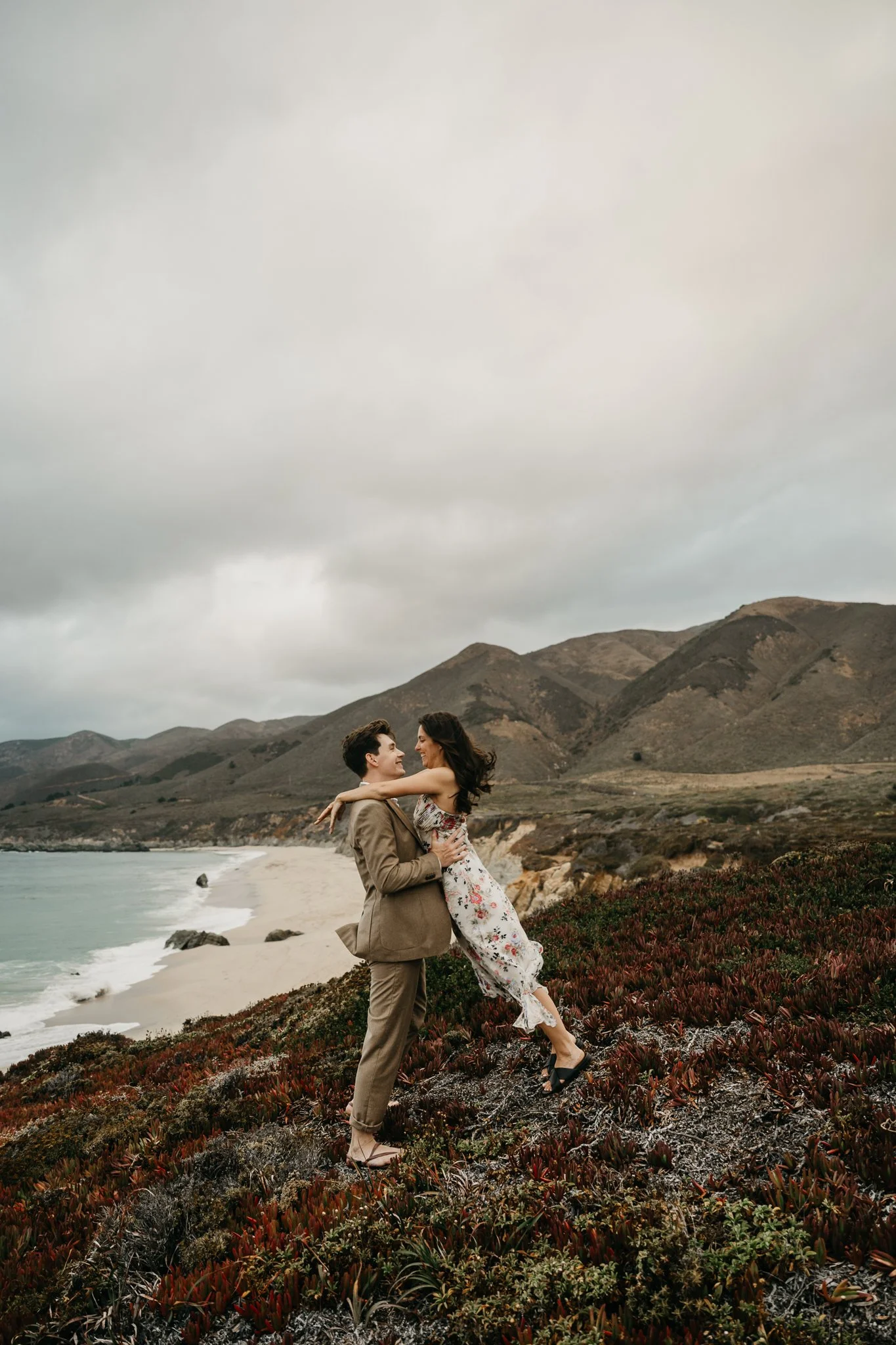 A couple dancing on a grassy hillside near a beach with cloudy skies and mountains in the background.
