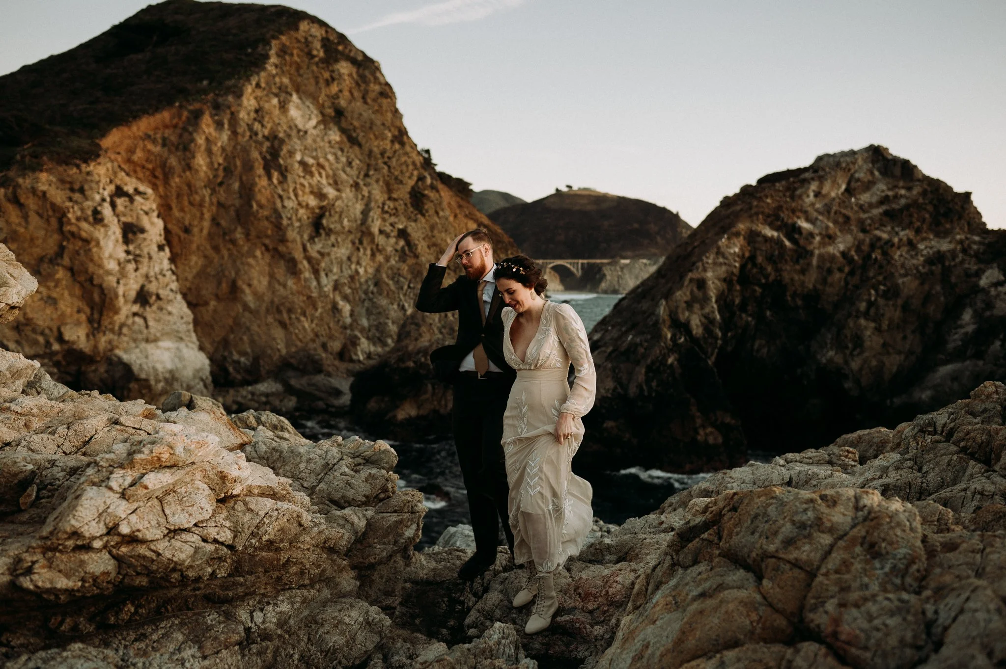 A couple in wedding attire standing on rocky terrain near the ocean, with large rocks and cliffs in the background, during sunset.