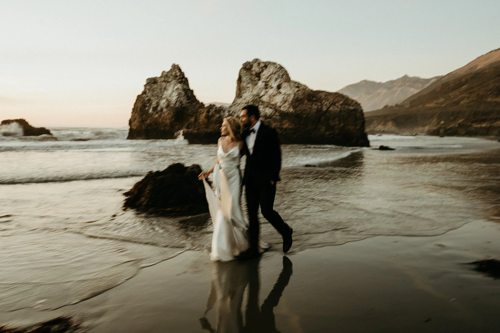 A bride and groom walking together on the beach near large rocks with mountains in the background at sunset.
