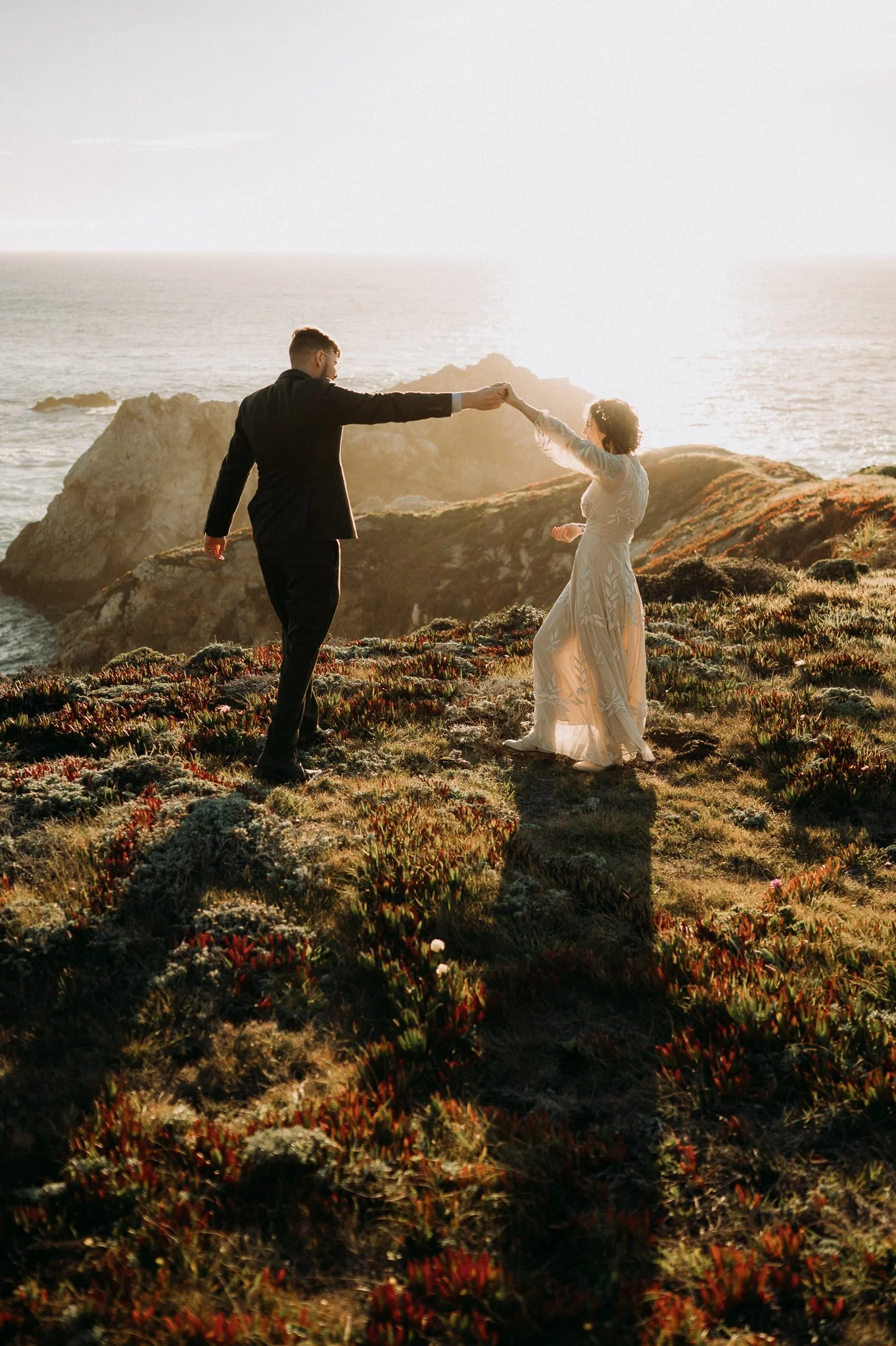 A couple in formal attire holding hands on a grassy coastal cliff at sunset, with ocean waves and rocky formations in the background.