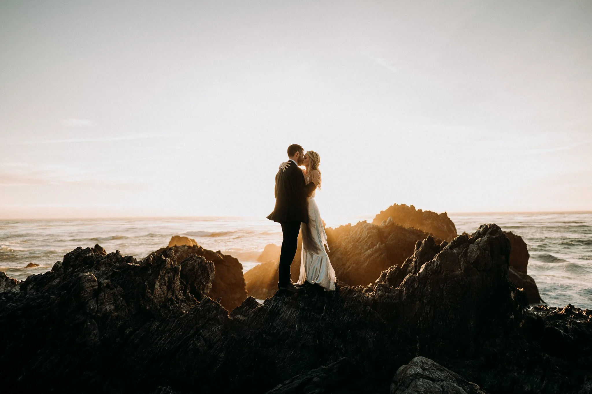 A couple in wedding attire sharing a kiss on rocks at the beach during sunset.