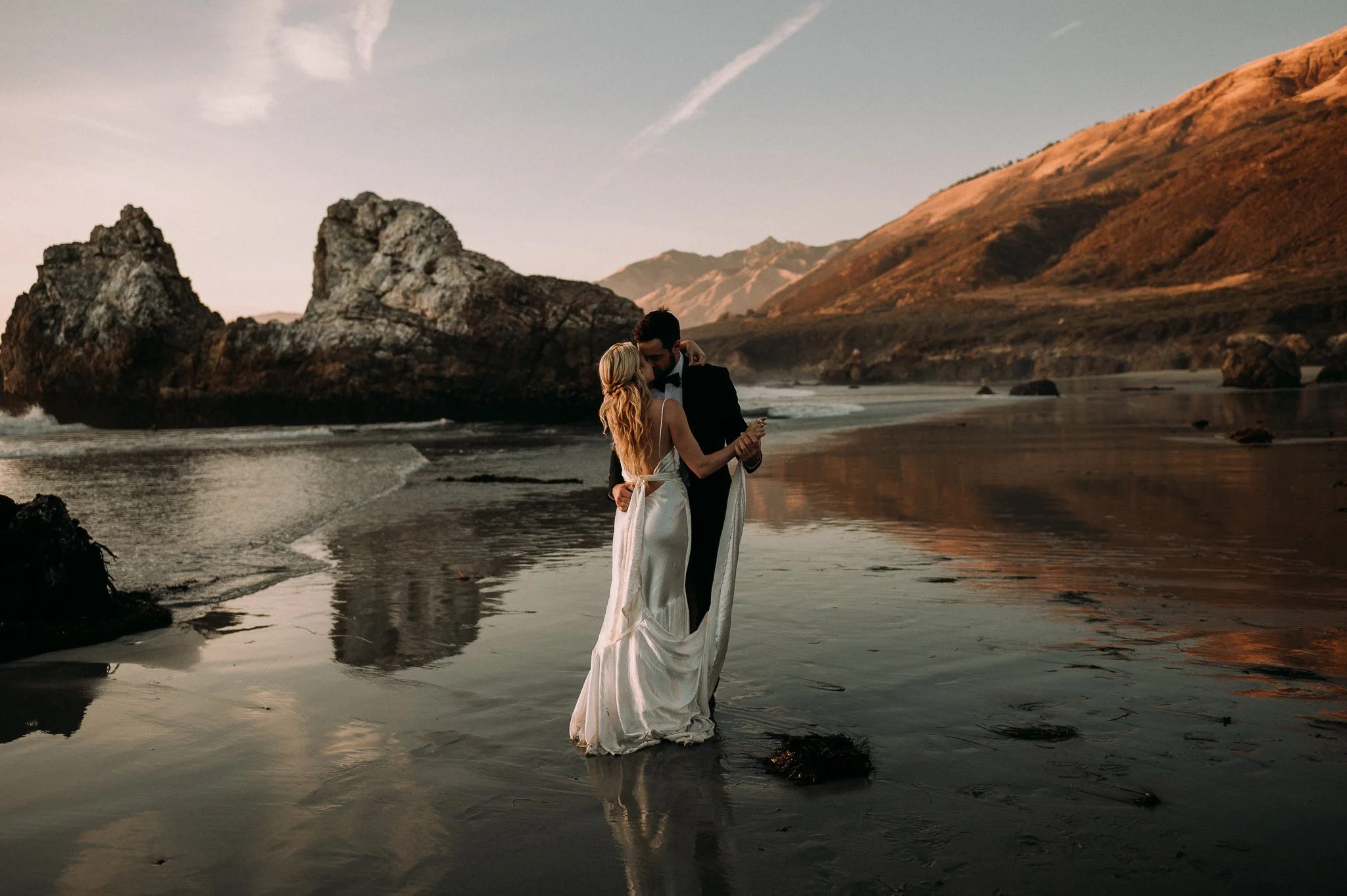 A couple in wedding attire standing on a beach at sunset, holding hands and embracing with rocky formations and mountains in the background.