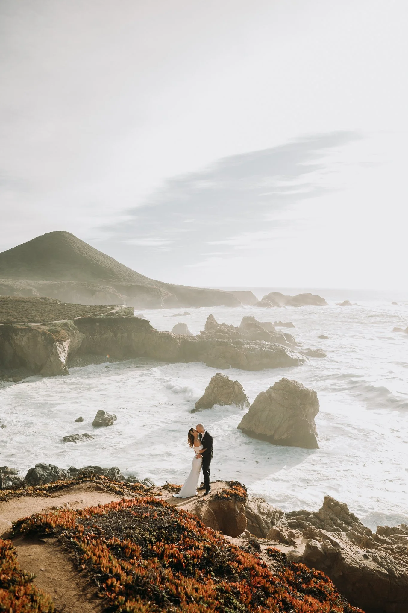 A couple dressed in wedding attire standing close to each other on a rocky cliff overlooking the ocean, with waves crashing against rocks and a hill in the background.