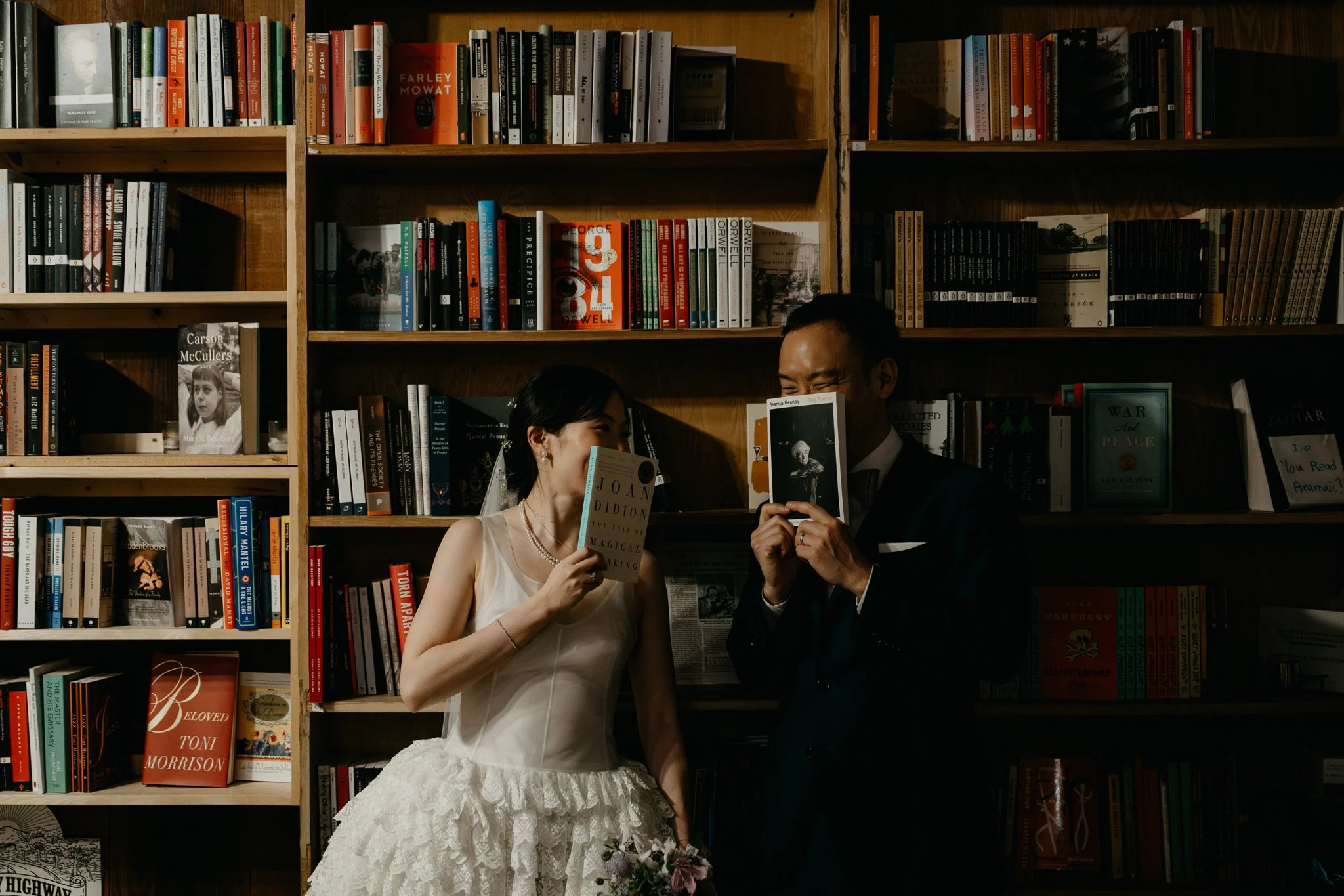 A bride and groom in wedding attire reading books and smiling in front of a large wooden bookshelf filled with books.