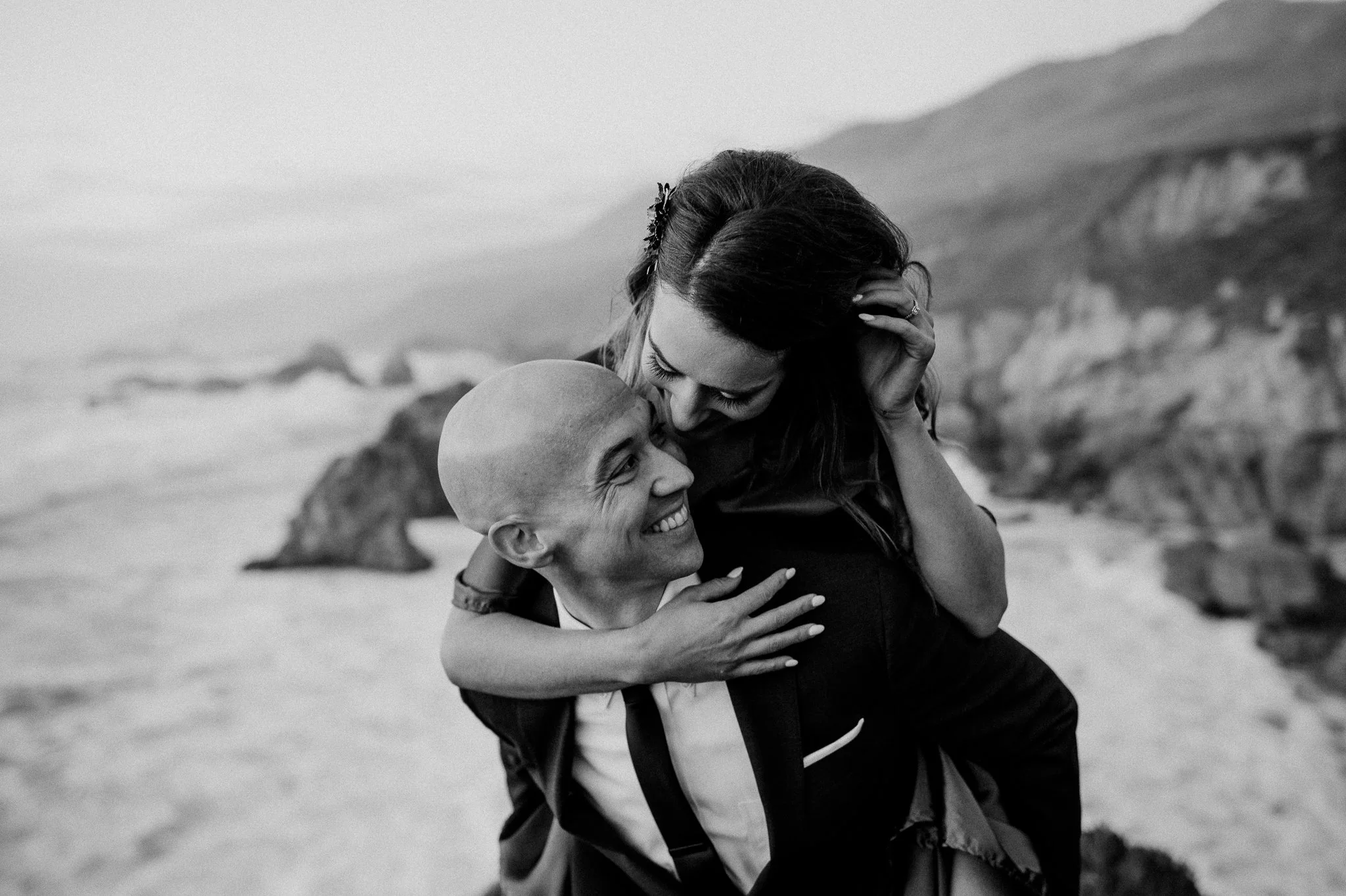 A black and white photo of a woman hugging and kissing a man with a bald head by the beach with cliffs in the background.