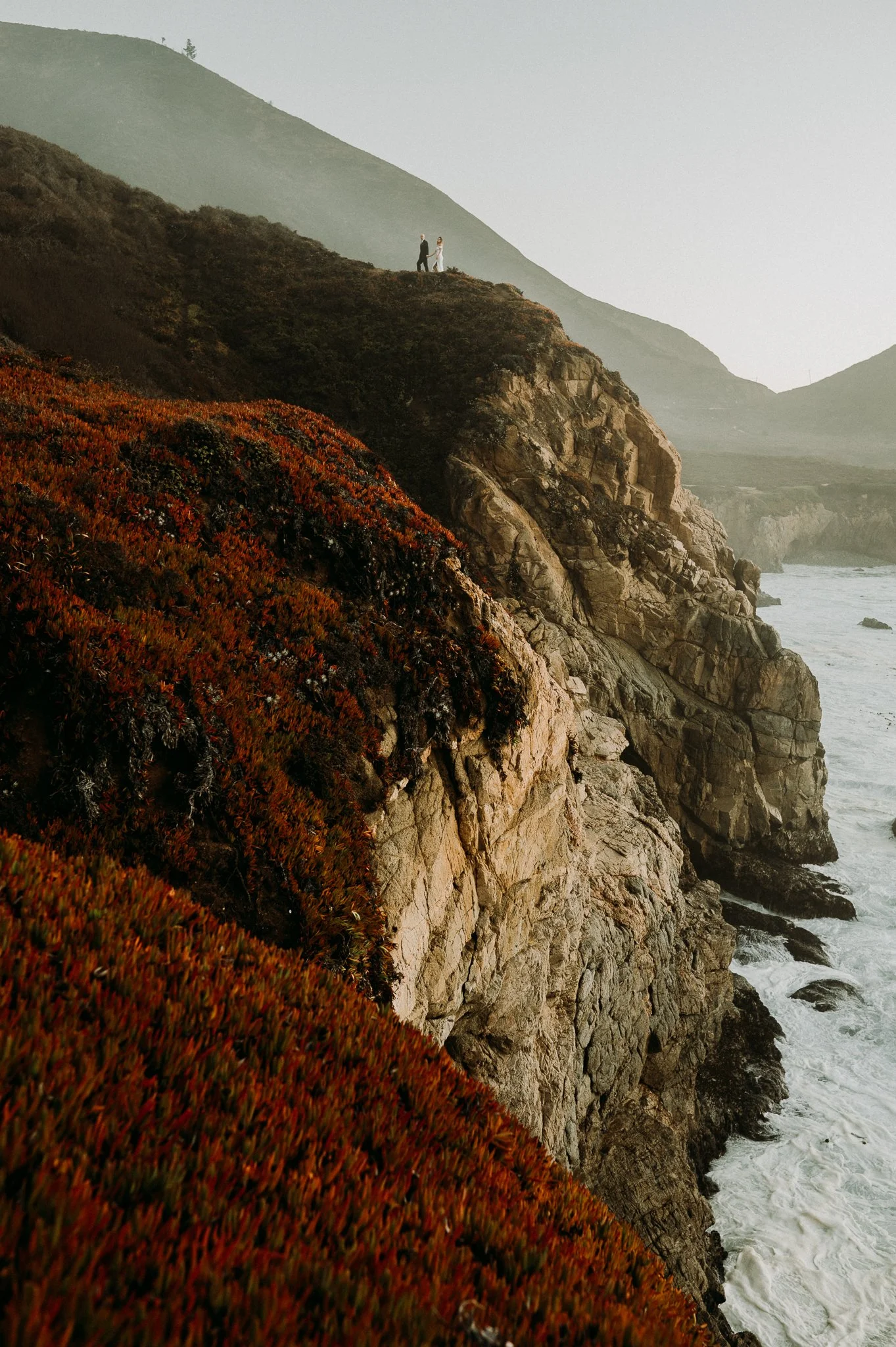 A couple holding hands standing on a cliff overlooking the ocean with rugged hills in the background.