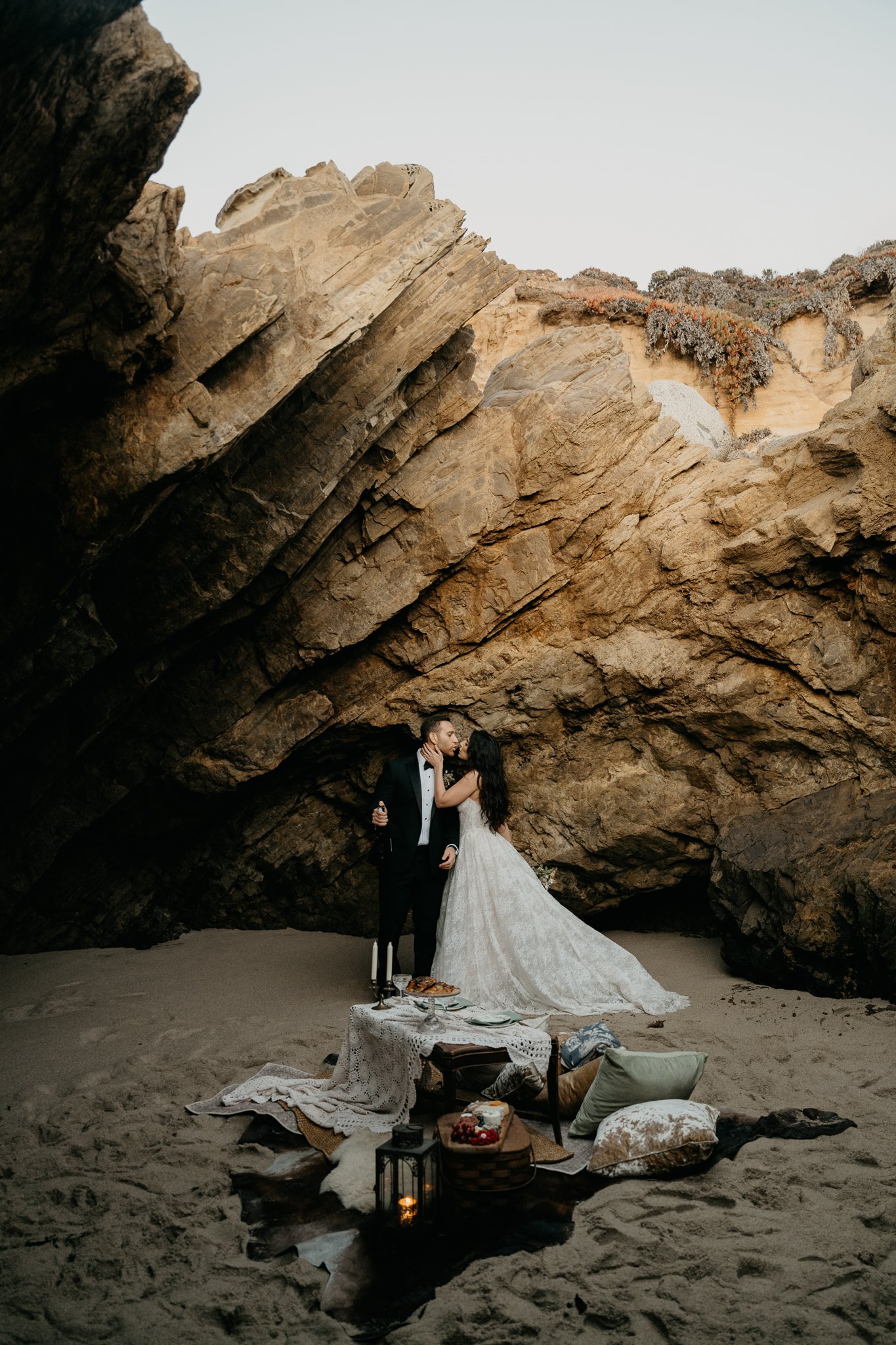 A couple dressed in wedding attire sharing a kiss under large rock formations on a sandy beach with a cozy picnic setup in front of them.