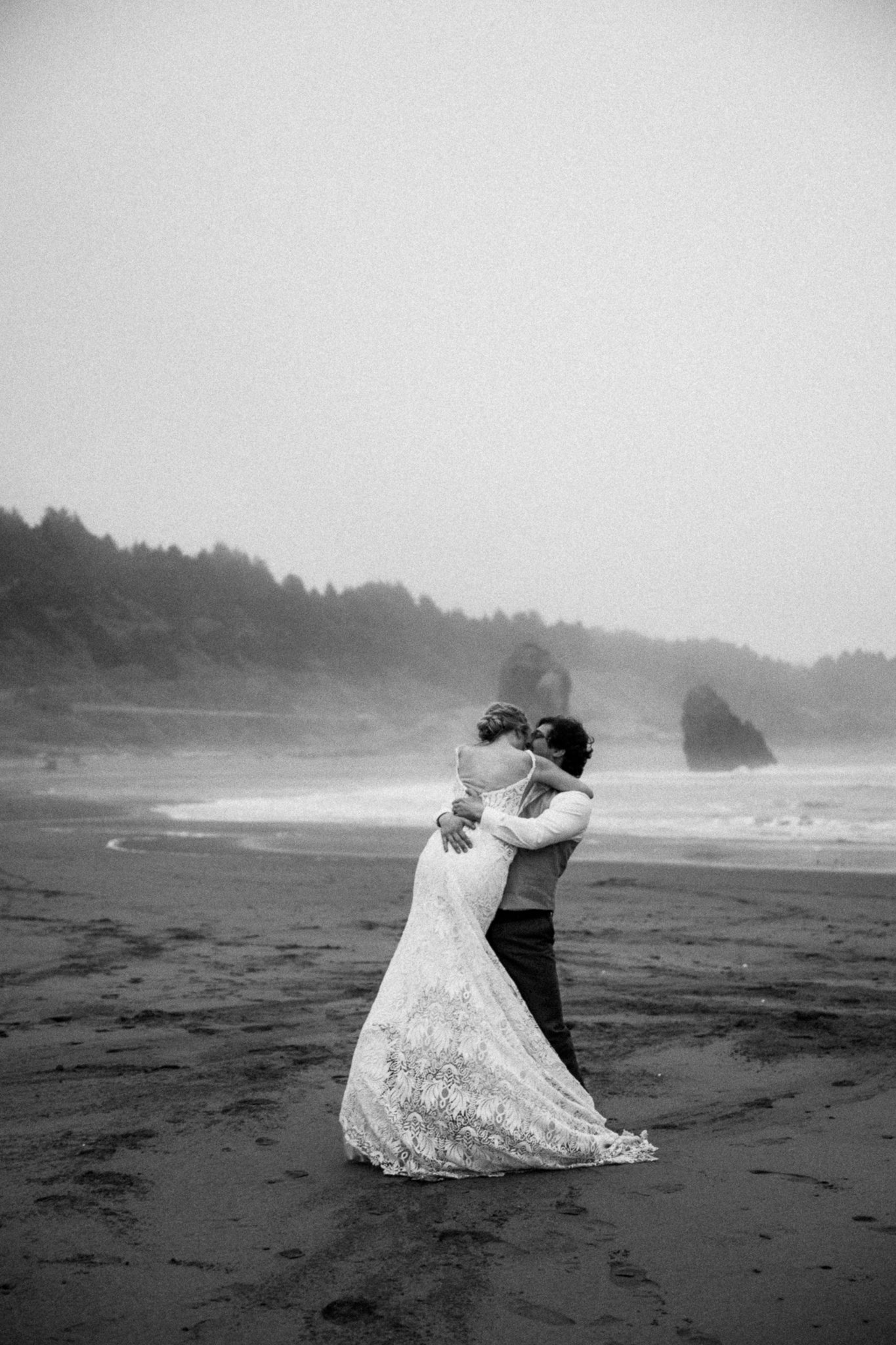 A black and white photograph of a couple sharing a kiss on a beach, with ocean waves and rocky formations in the background.