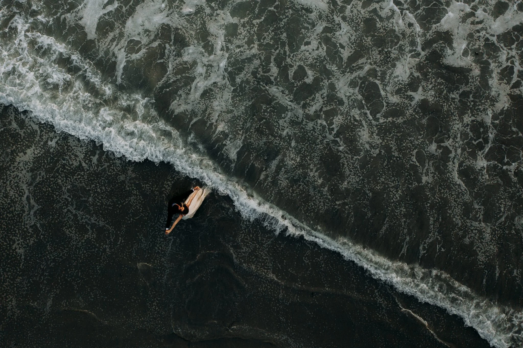 A person in a white dress lying on a surfboard in the ocean, with dark water and white foamy waves.