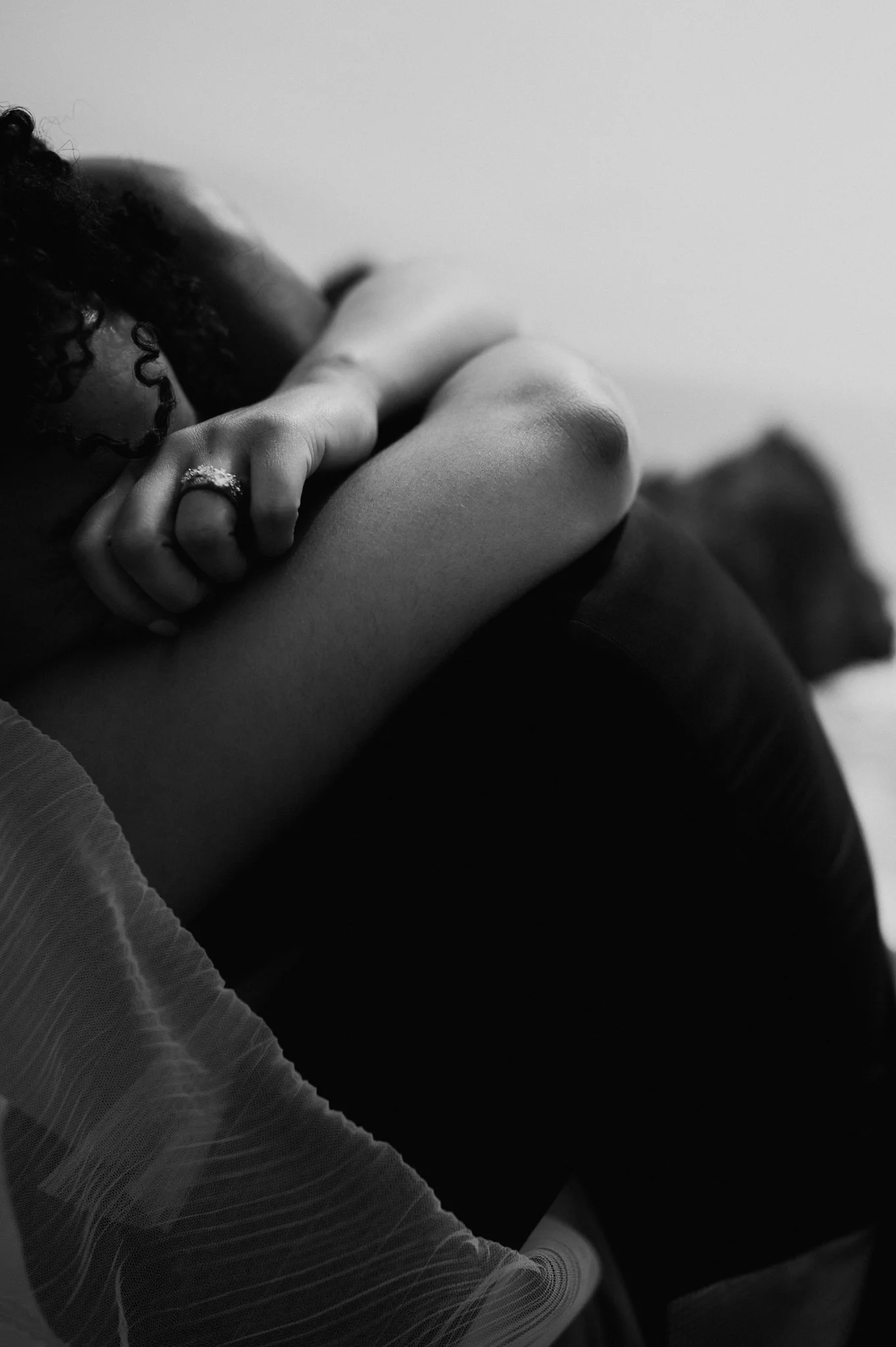Close-up black and white photo of a woman with curly hair, lying on her side with her hand resting on her knee. She is wearing a ring on her finger and a sheer, textured top.