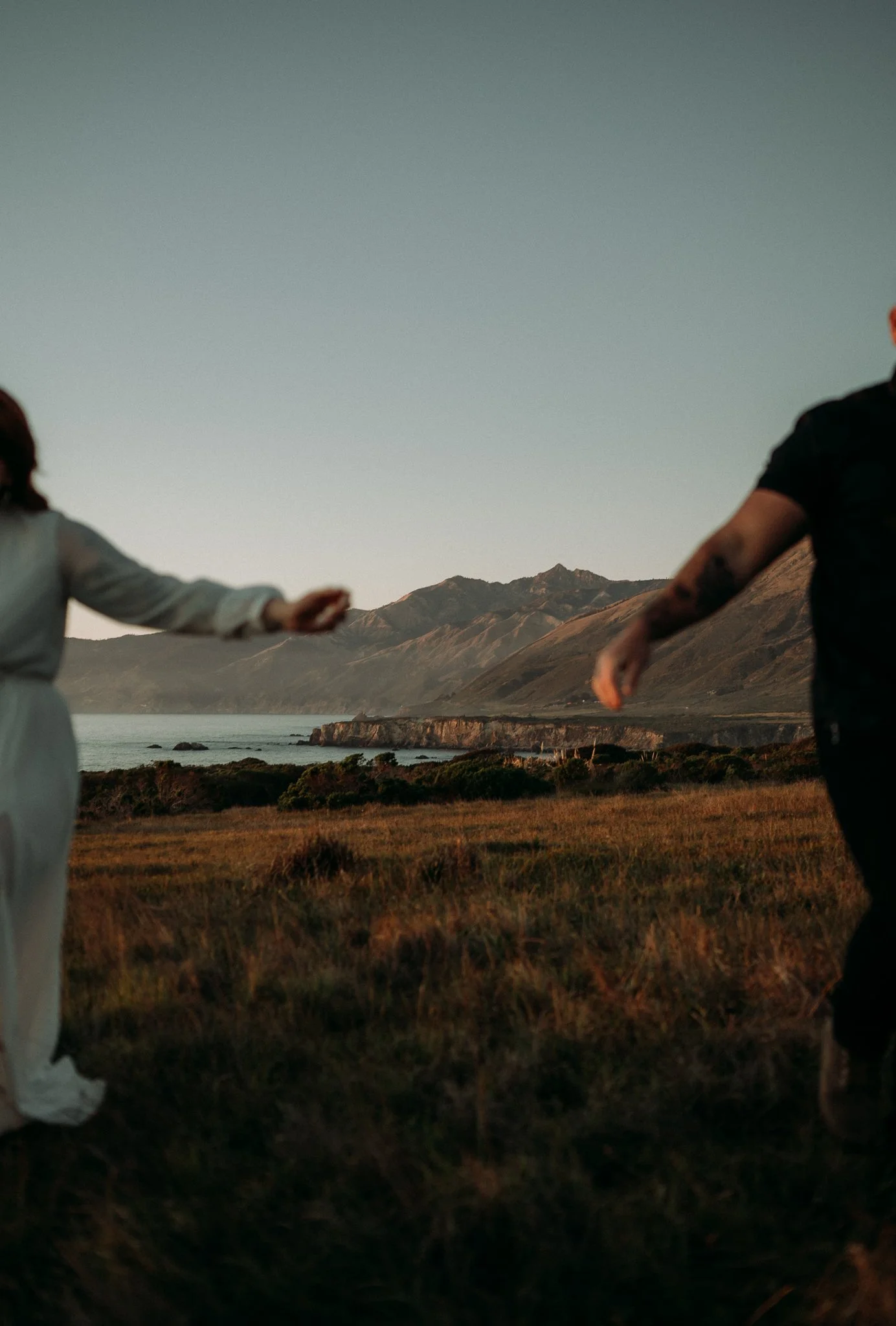 Couple holding hands outdoors at sunset near a coastal landscape with mountains in the background.