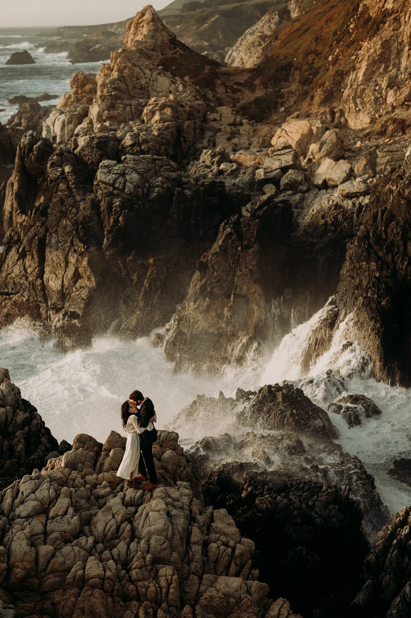 A couple stands on rocky cliffs by the ocean, embracing each other as waves crash against the rocks.