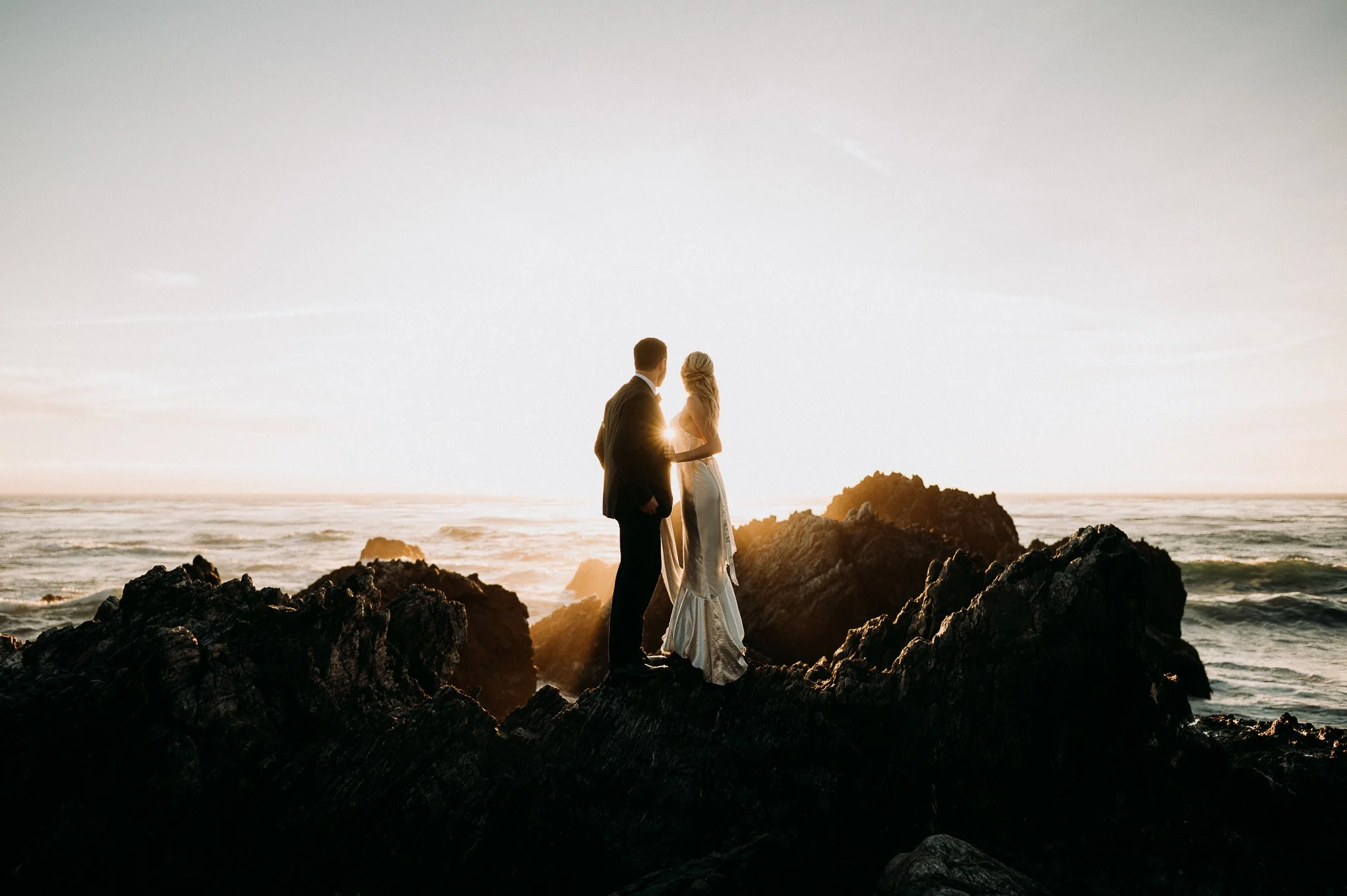 A couple standing on rocks by the ocean at sunset, facing each other and holding hands.