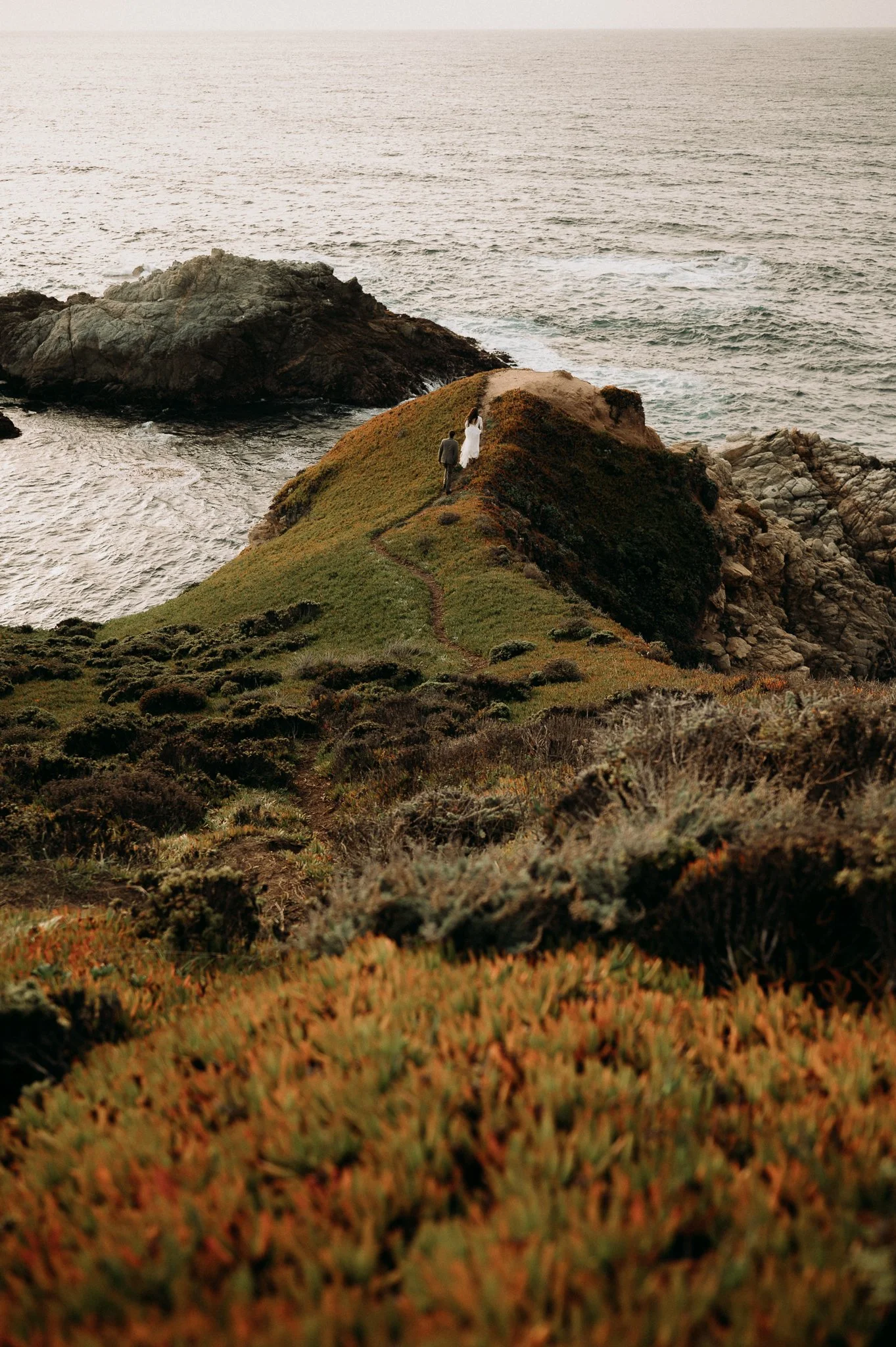 A couple walking on a narrow trail along a grassy coastal hillside towards the ocean at sunset.