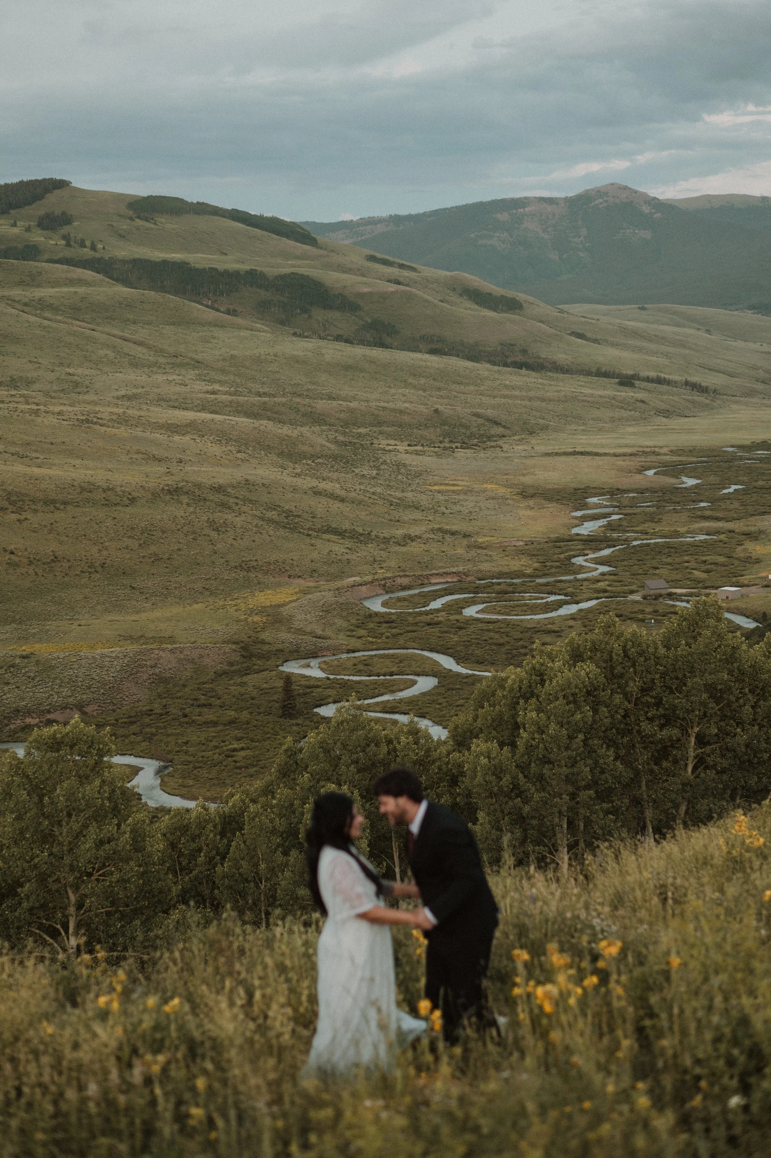 A couple holding hands and smiling at each other in a field of yellow flowers with rolling hills and a winding river in the background.