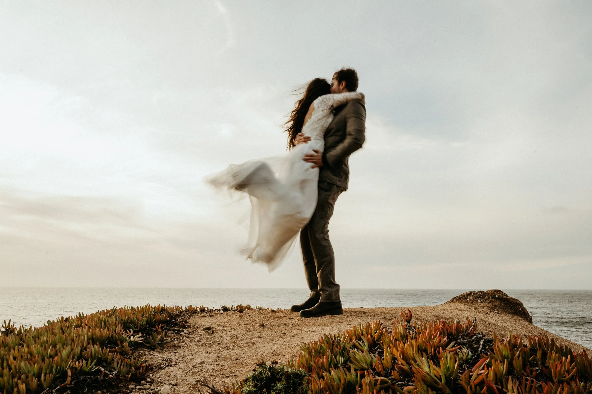 A couple embracing on a rocky cliff near the ocean, with the woman wearing a white dress and the man wearing a jacket, against a cloudy sky.