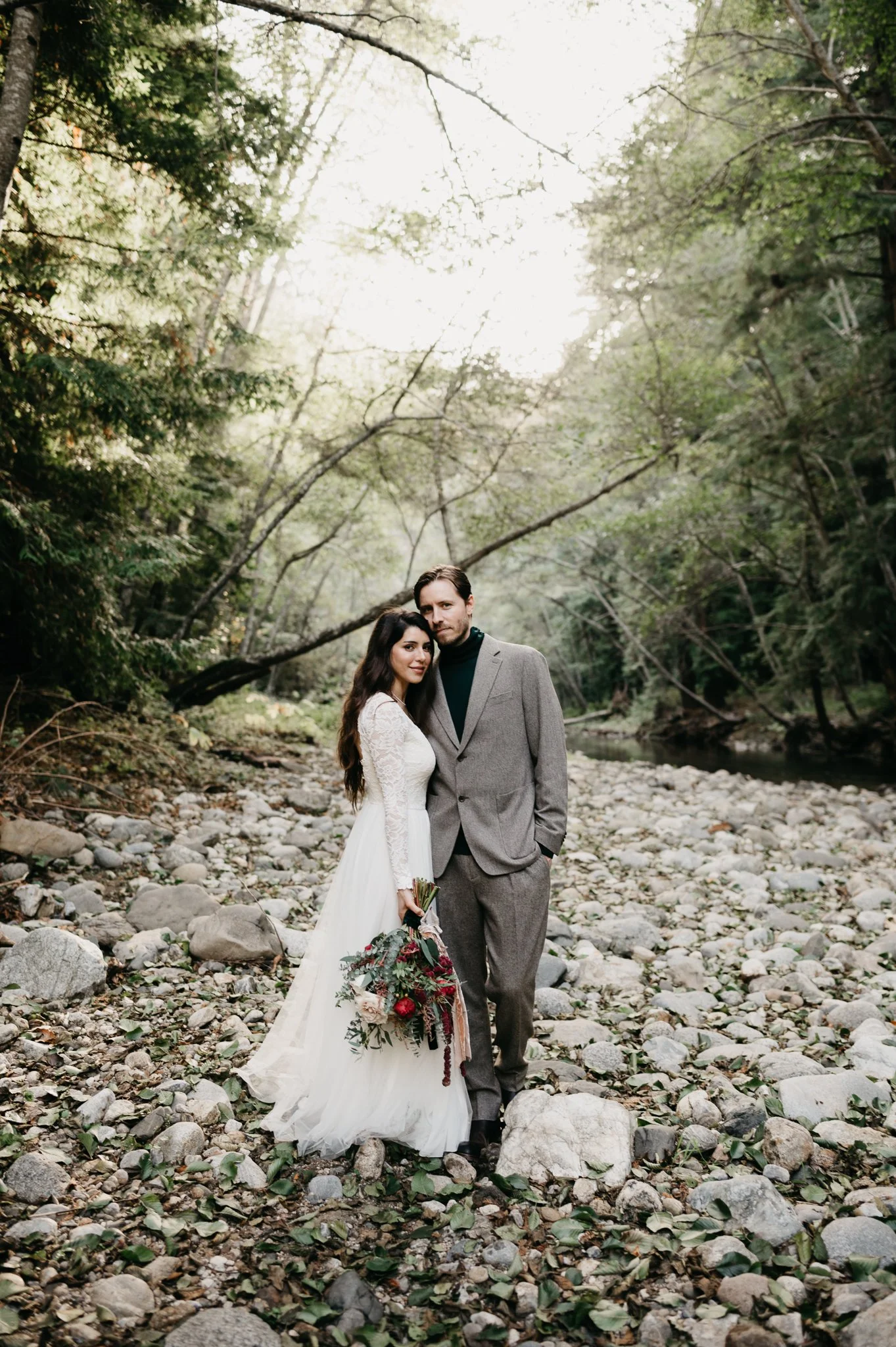 A bride and groom standing together on a rocky riverbed surrounded by trees and greenery, with sunlight filtering through the branches.