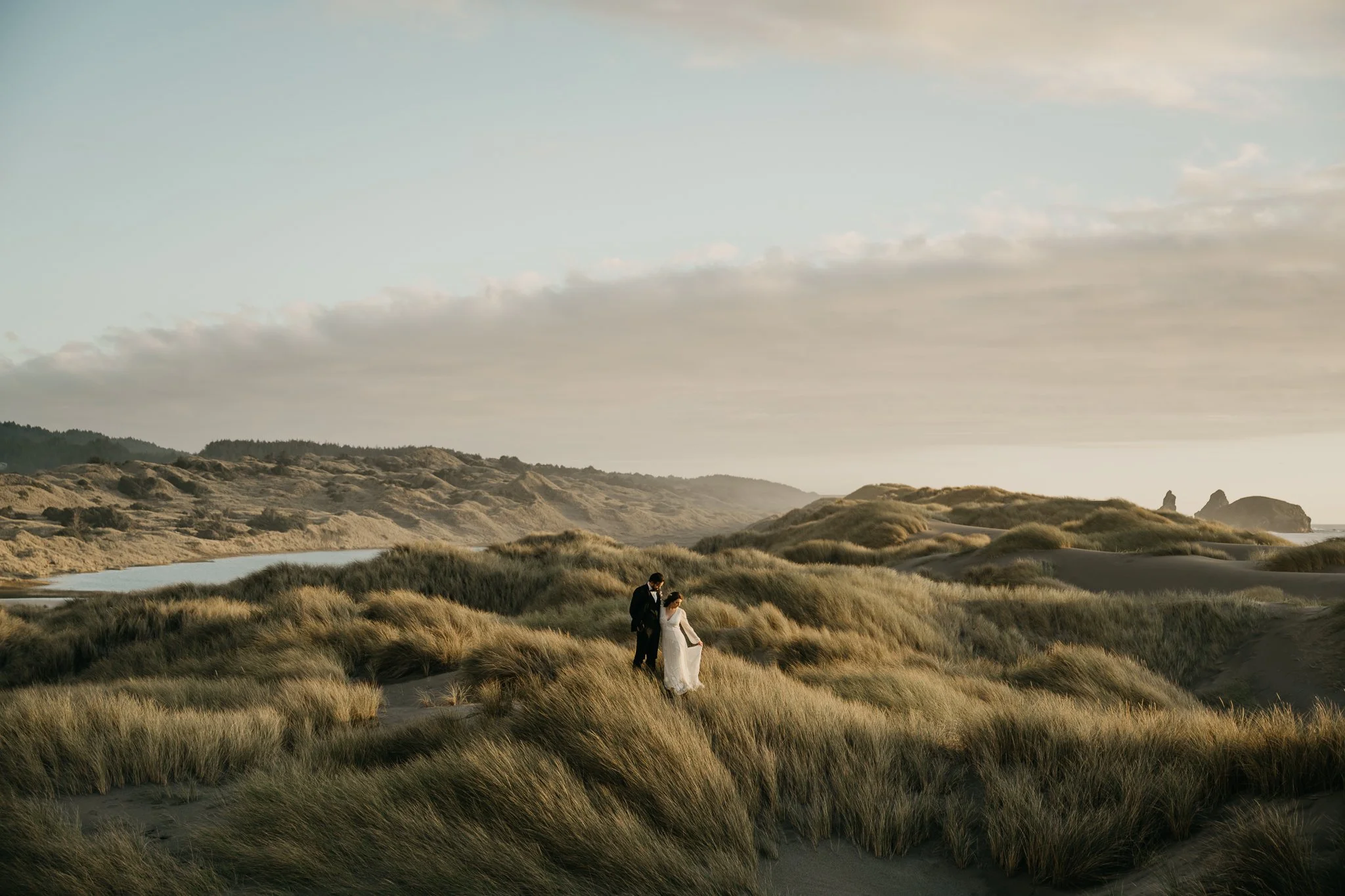 A couple in wedding attire walking through sandy dunes with tall grass on a beach at sunset.