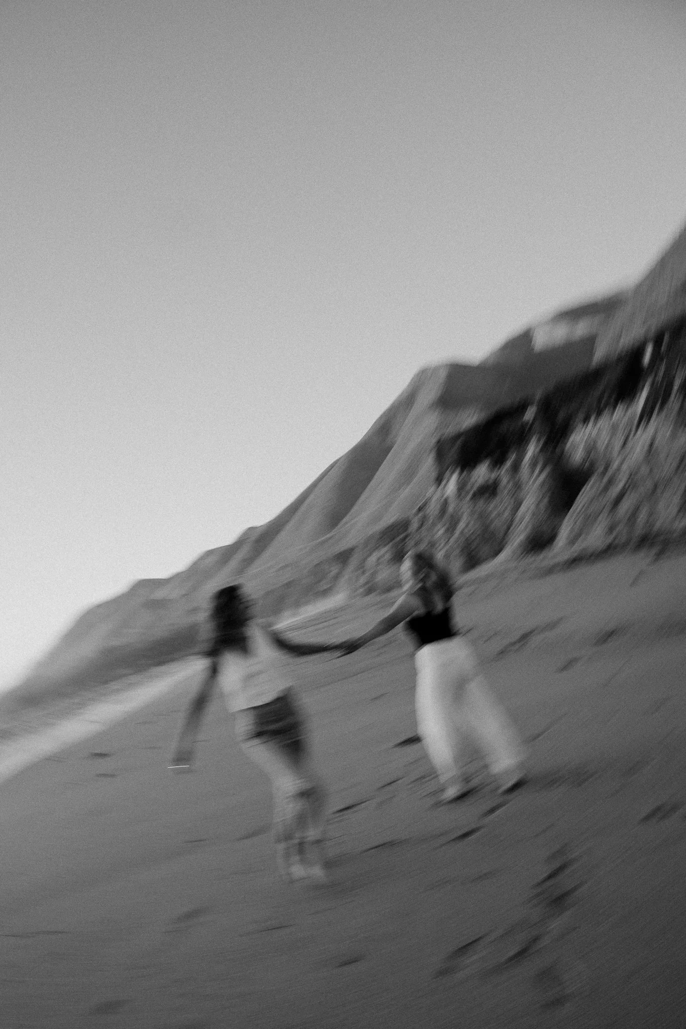 Two women holding hands and walking on a beach, with cliffs in the background, black and white photo.
