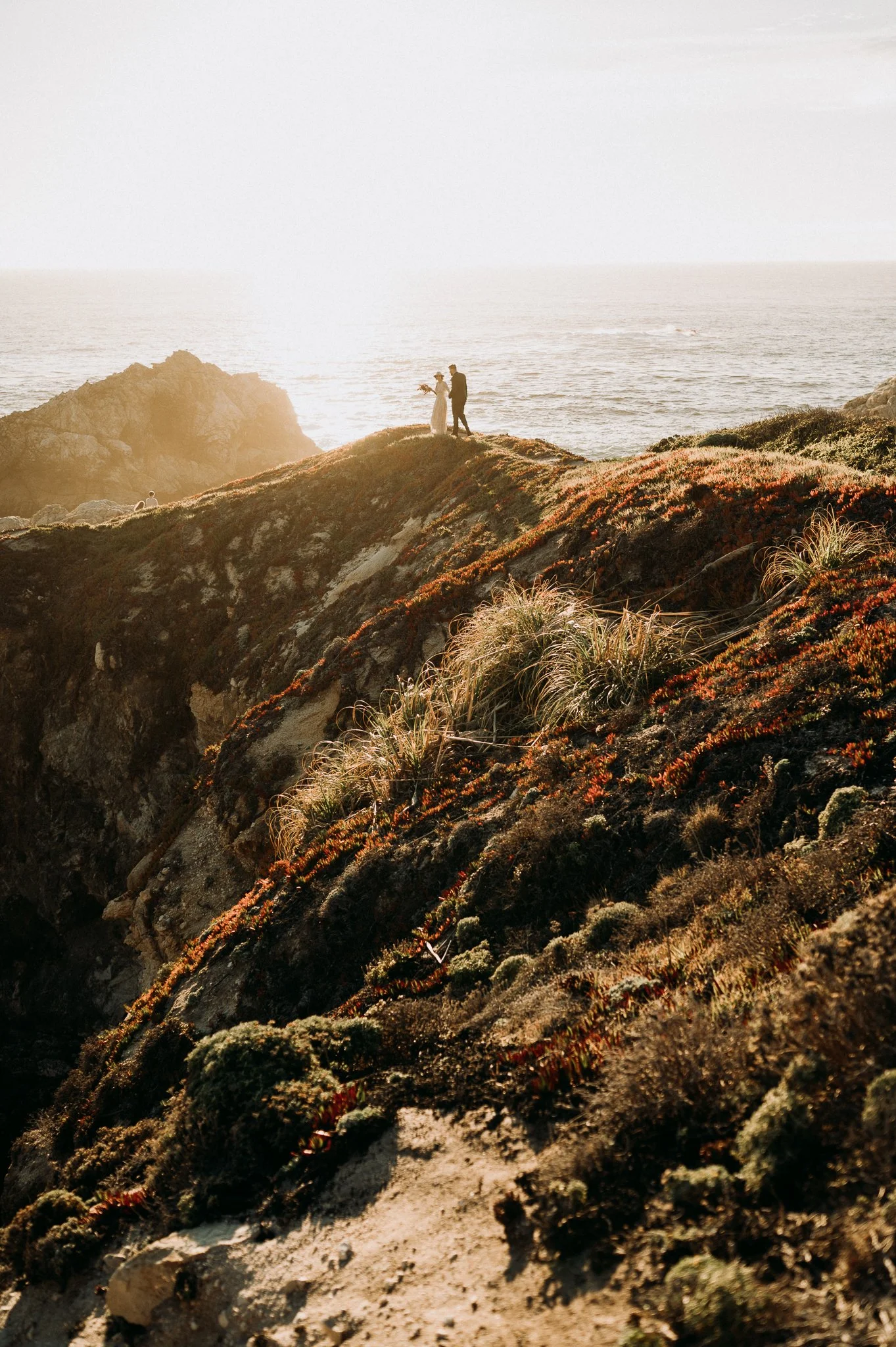 A couple standing on a grassy cliff overlooking the ocean at sunset, with rocky formations in the background.