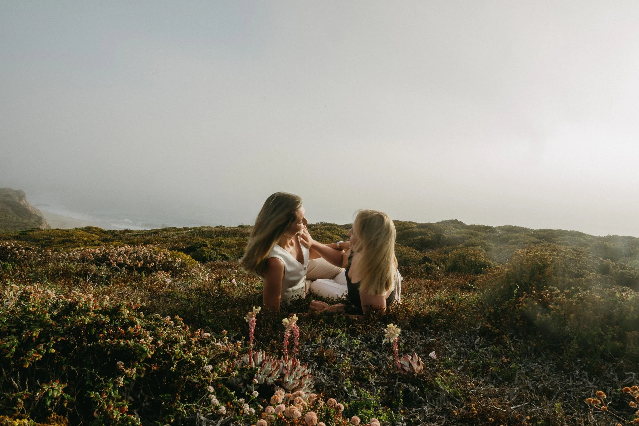 Two women sitting amidst flowering plants and bushes, facing each other, with the ocean and a cloudy sky in the background.