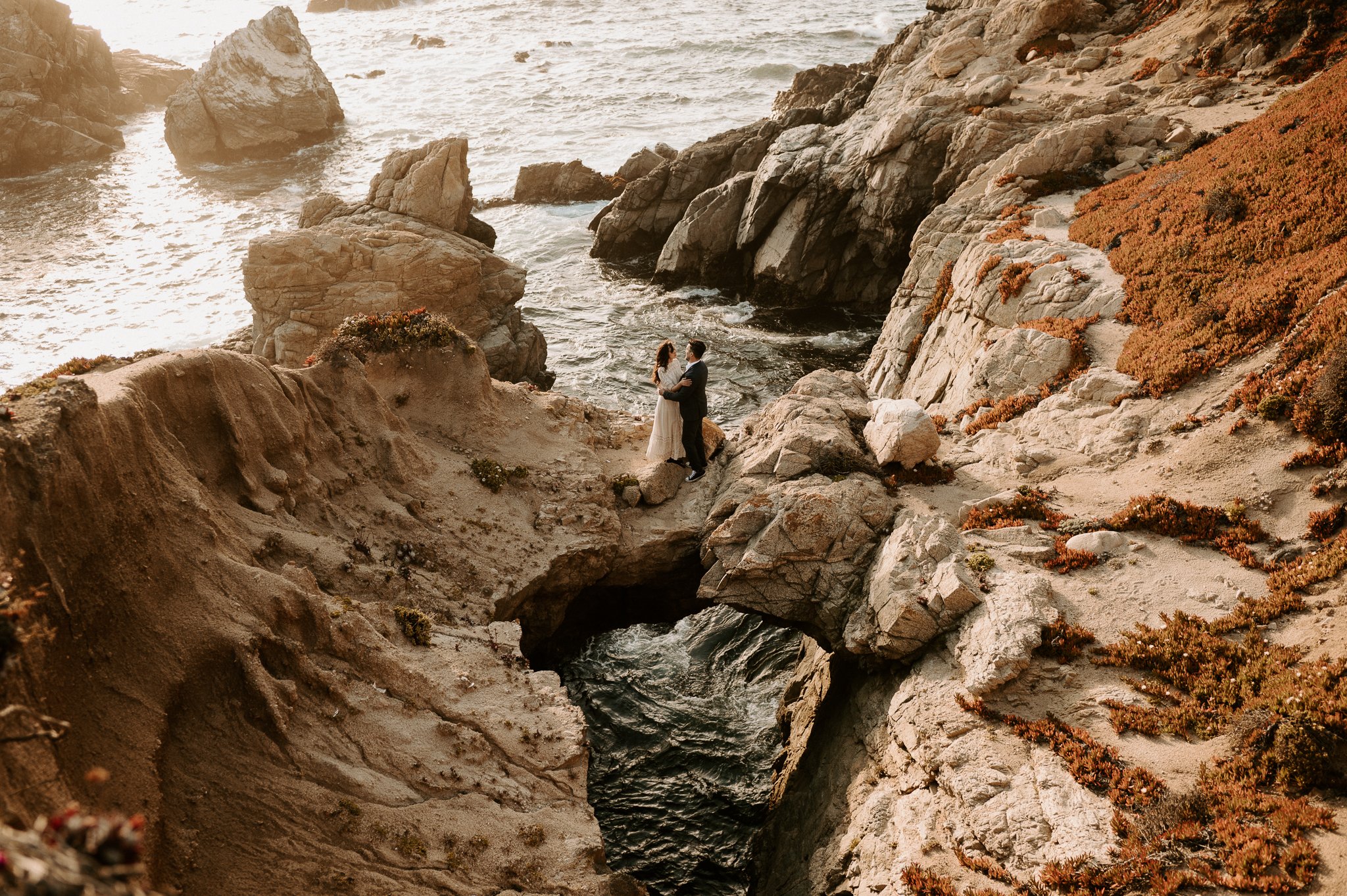 A couple stands on rocky coastal terrain, embracing with the ocean in the background during sunset, with waves hitting the shore and reddish-brown coastal vegetation.