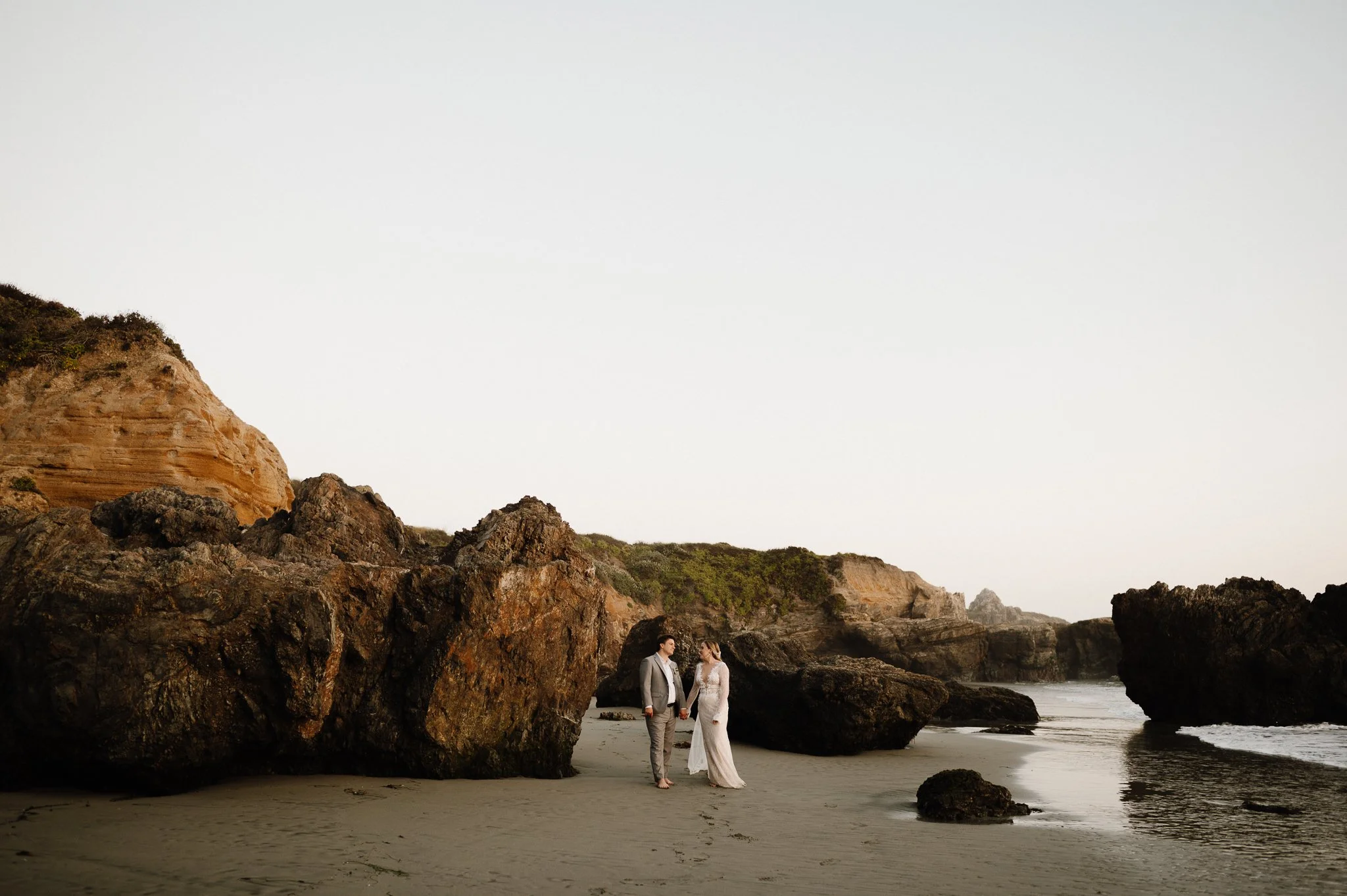 A bride and groom holding hands on a beach with large rocks and cliffs nearby at sunset.